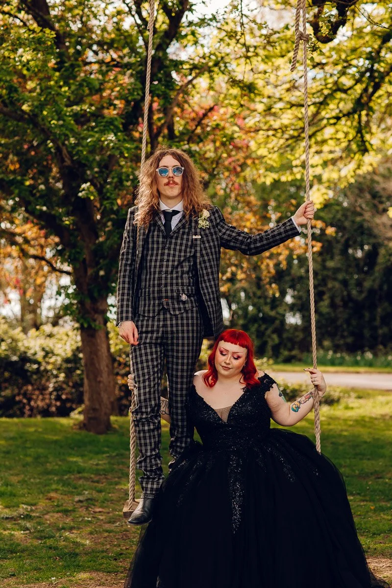 A bride in a black dress sits on a swing whilst her husband stands on the swing casually