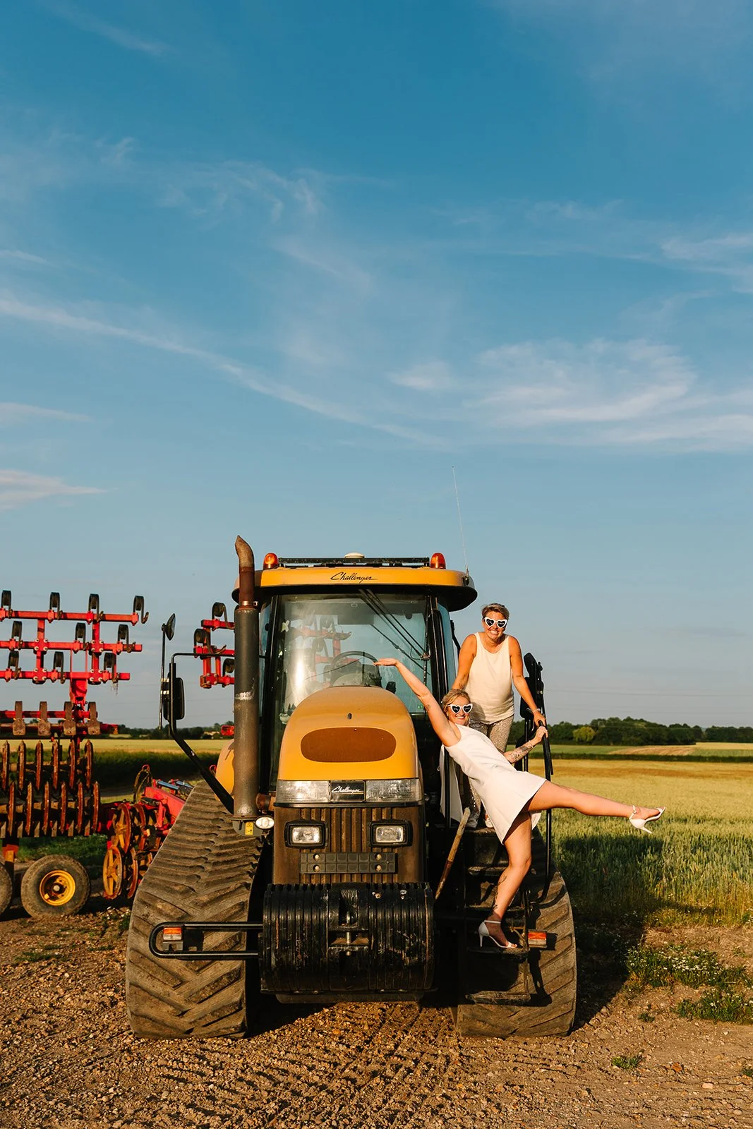 Two women wearing white dresses and sunglasses posing on a large yellow tractor in a field during sunset.
