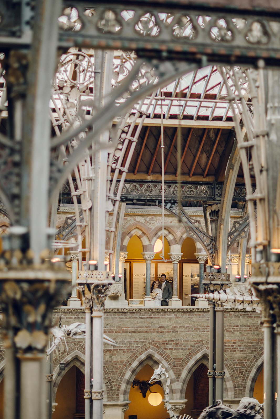 Inside a museum with architectural details, a couple stands on an upper level in front of informational panels, surrounded by skeletal displays and iron framework.