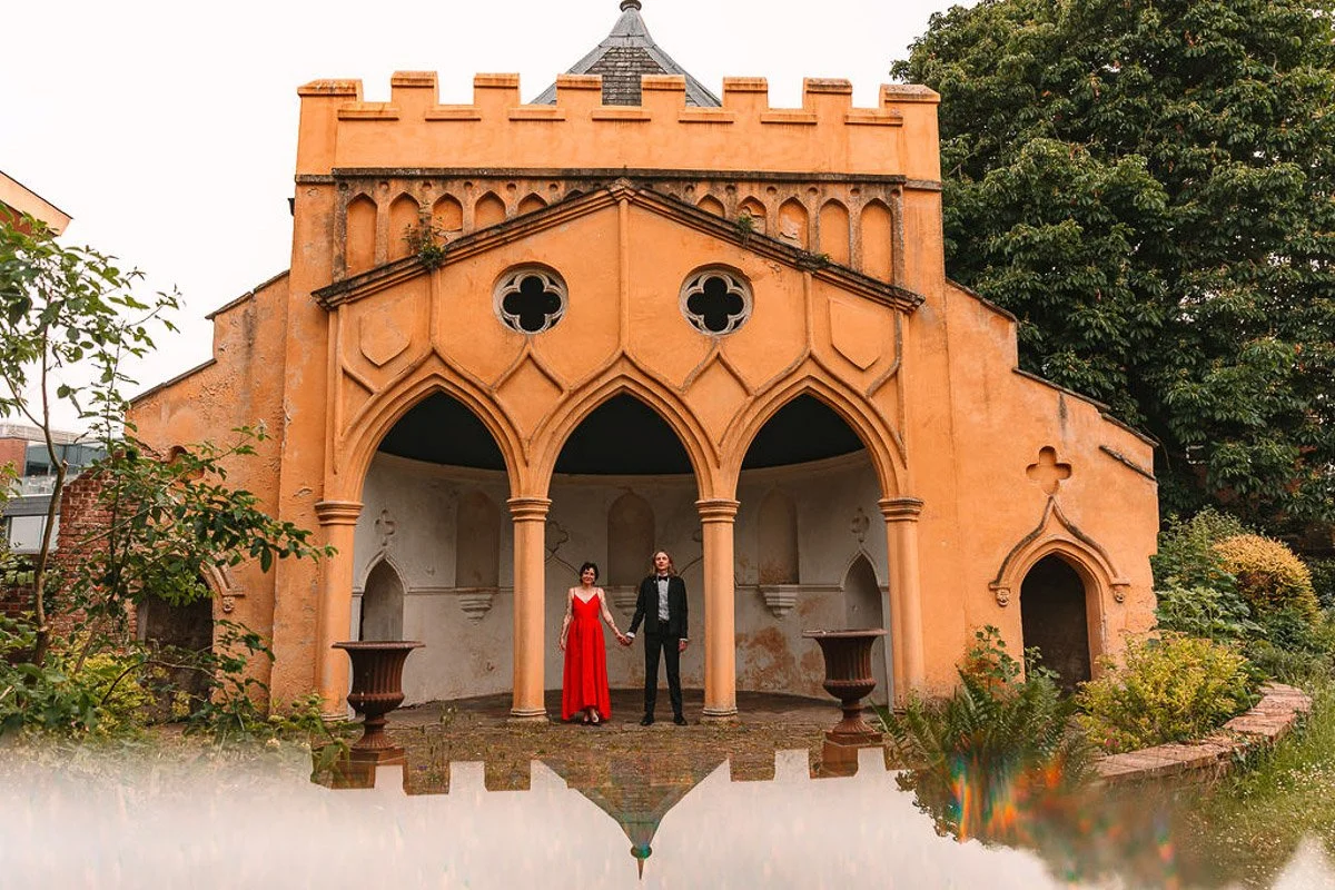 A bride in a red dress with short black hair holds hands with her groom in a black tuxedo. They stand in an arch of a gothic building outside.