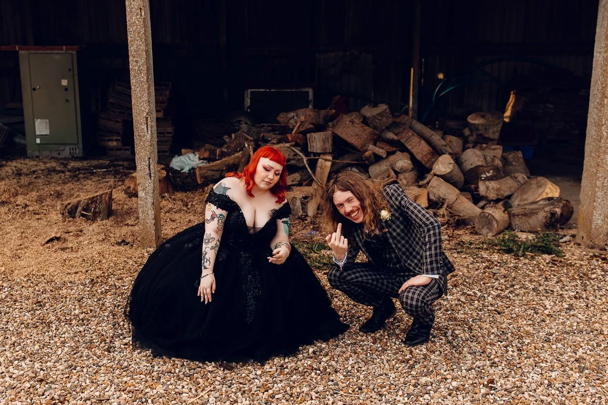 A fun image of the bride in a black dress and a groom holding his fingers up against an industrial backdrop at houchins wedding venue
