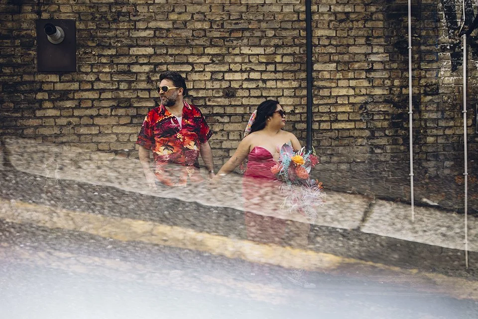 A man and woman holding hands, dressed in colorful clothing, standing against a brick wall with a black speaker and piping, with a blurred foreground.