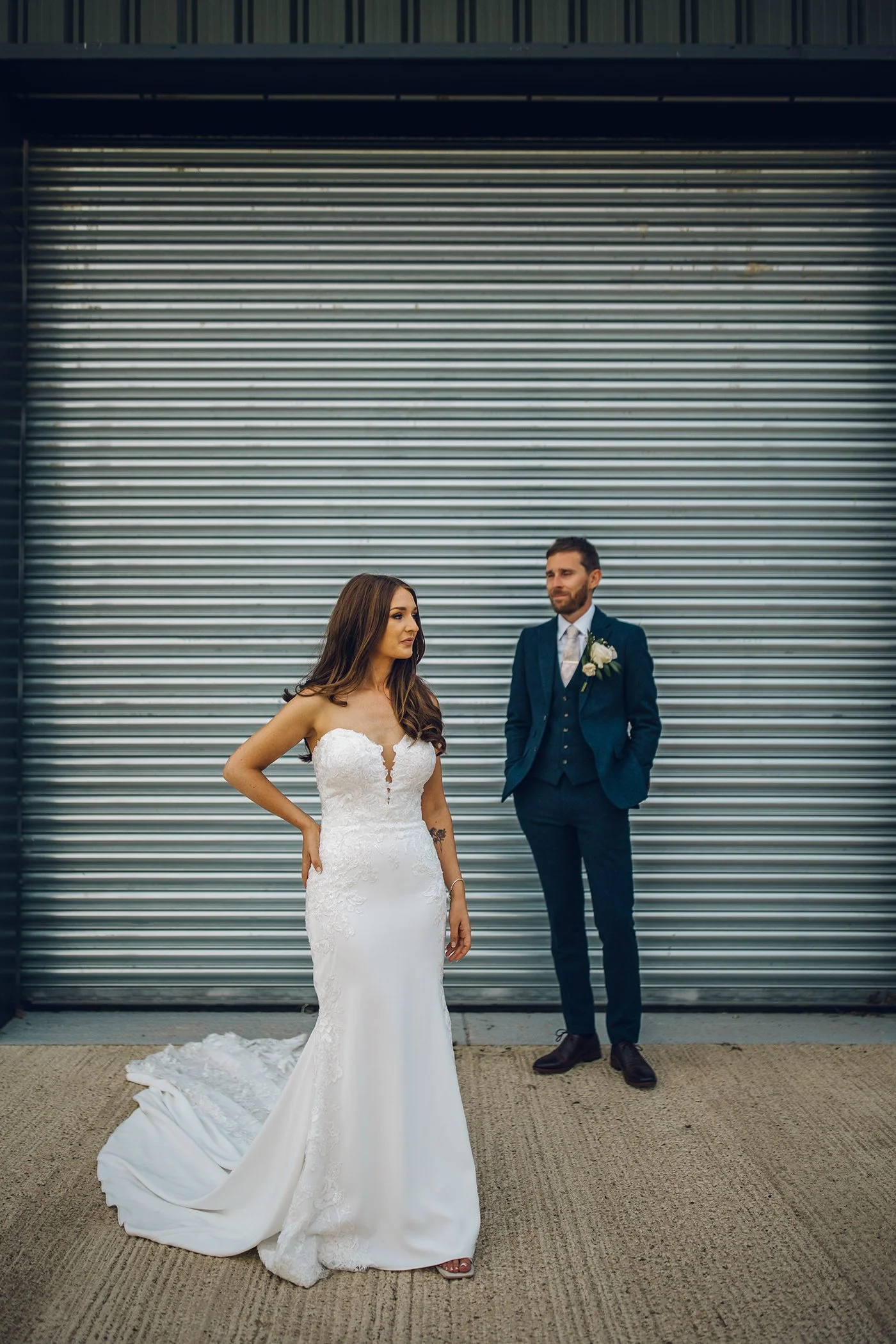 A bride in a white wedding dress and a groom in a blue suit standing outside in front of a closed metal shutter.