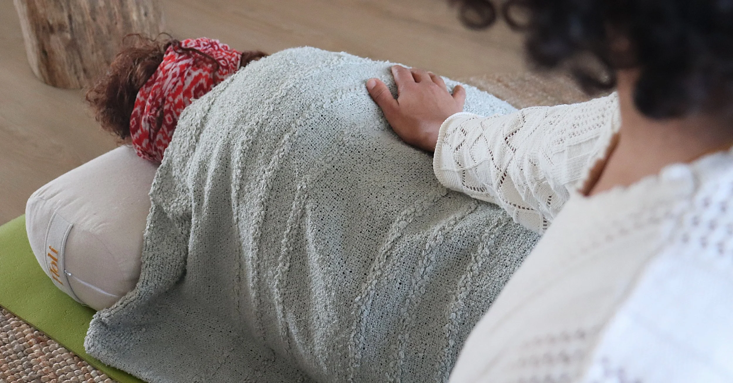 Persoon ligt op een relaxmat met een warme deken, terwijl iemand anders hun hand op hun rug legt in een ontspannings- of massage of yogatherapie setting