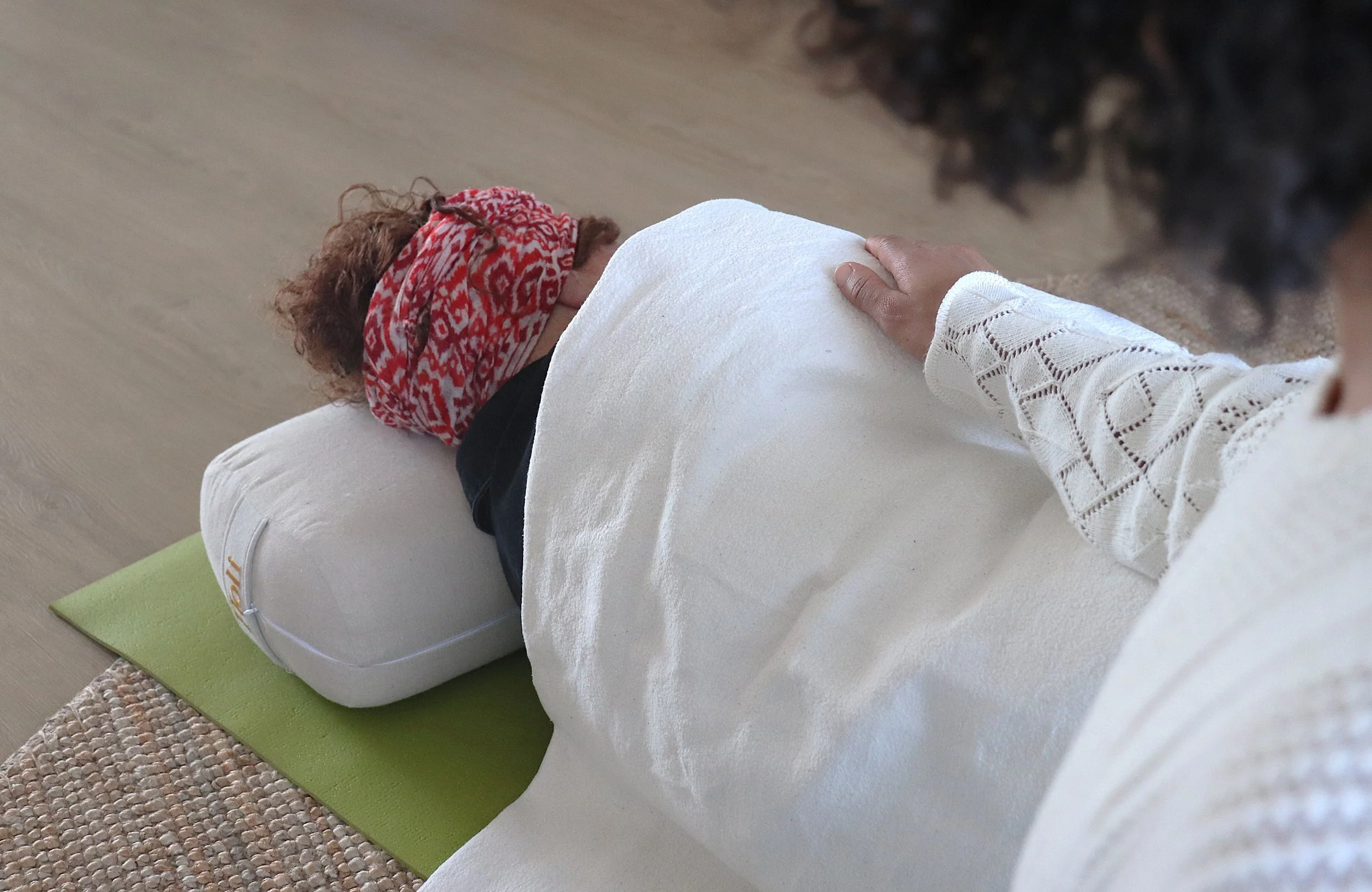 Persoon ligt op een matras met een kussen onder het hoofd, terwijl een andere persoon met een hand op de rug de persoon verzorgt, klaar voor een yogatherapie of reflexologie behandeling