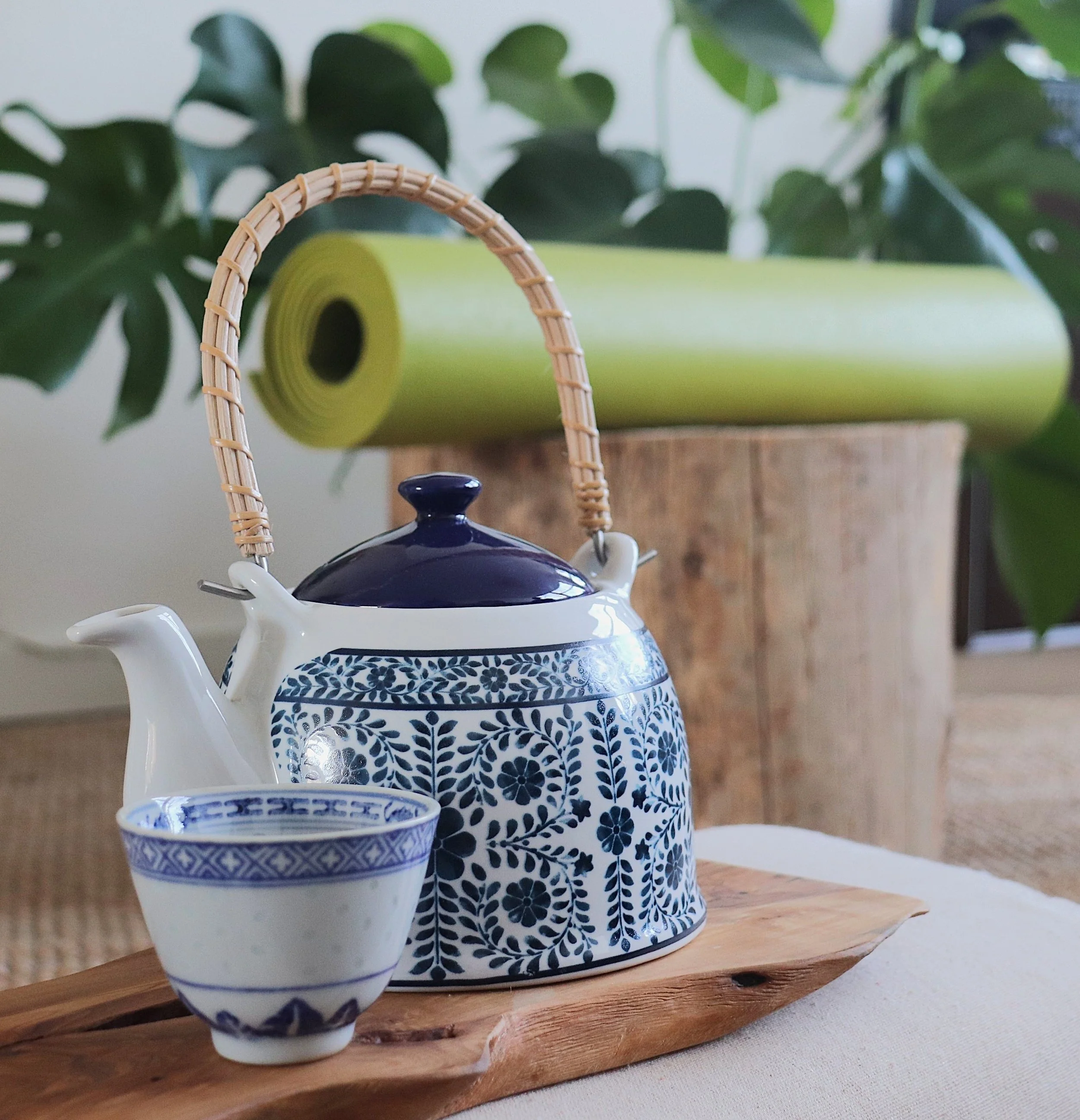 Een blauwe en witte keramiek theepot met een bloemmotief, een bijpassende kop, een bamboe mand met een handvat en een groene yoga mat op een houten plank, een plant op de achtergrond.