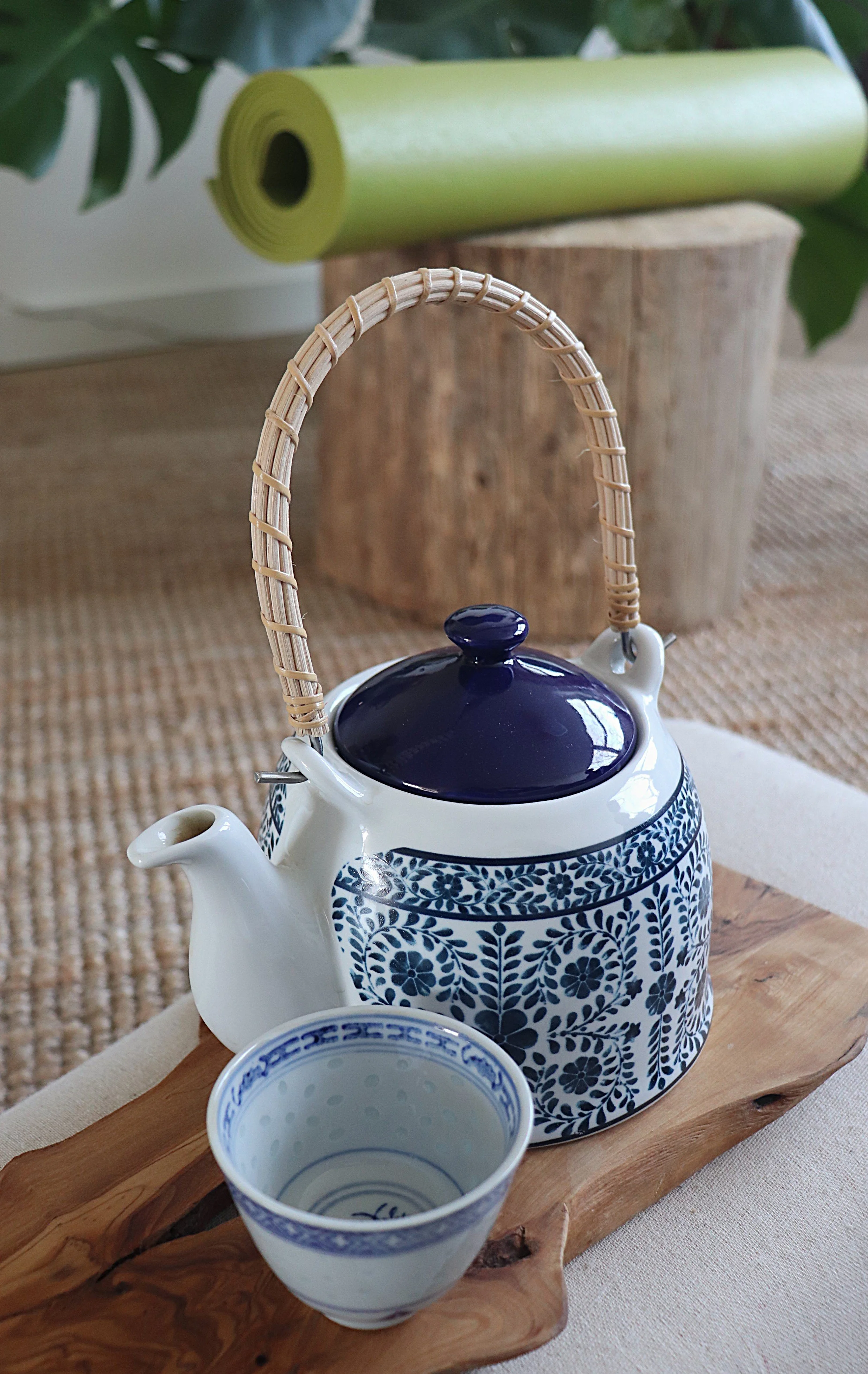 Een keramieke theepot met blauwe bloemmotief en een bijpassende kop, geplaatst op een houten snijbord, met een yogamat en yogablokken op de achtergrond