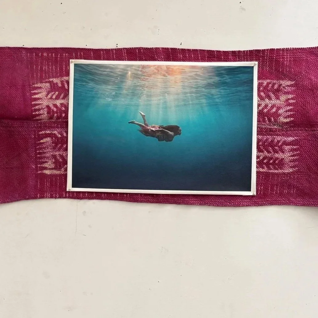 Photograph of a woman swimming underwater, displayed on a pink cloth with white patterns against a white wall. Vanuatu water music.