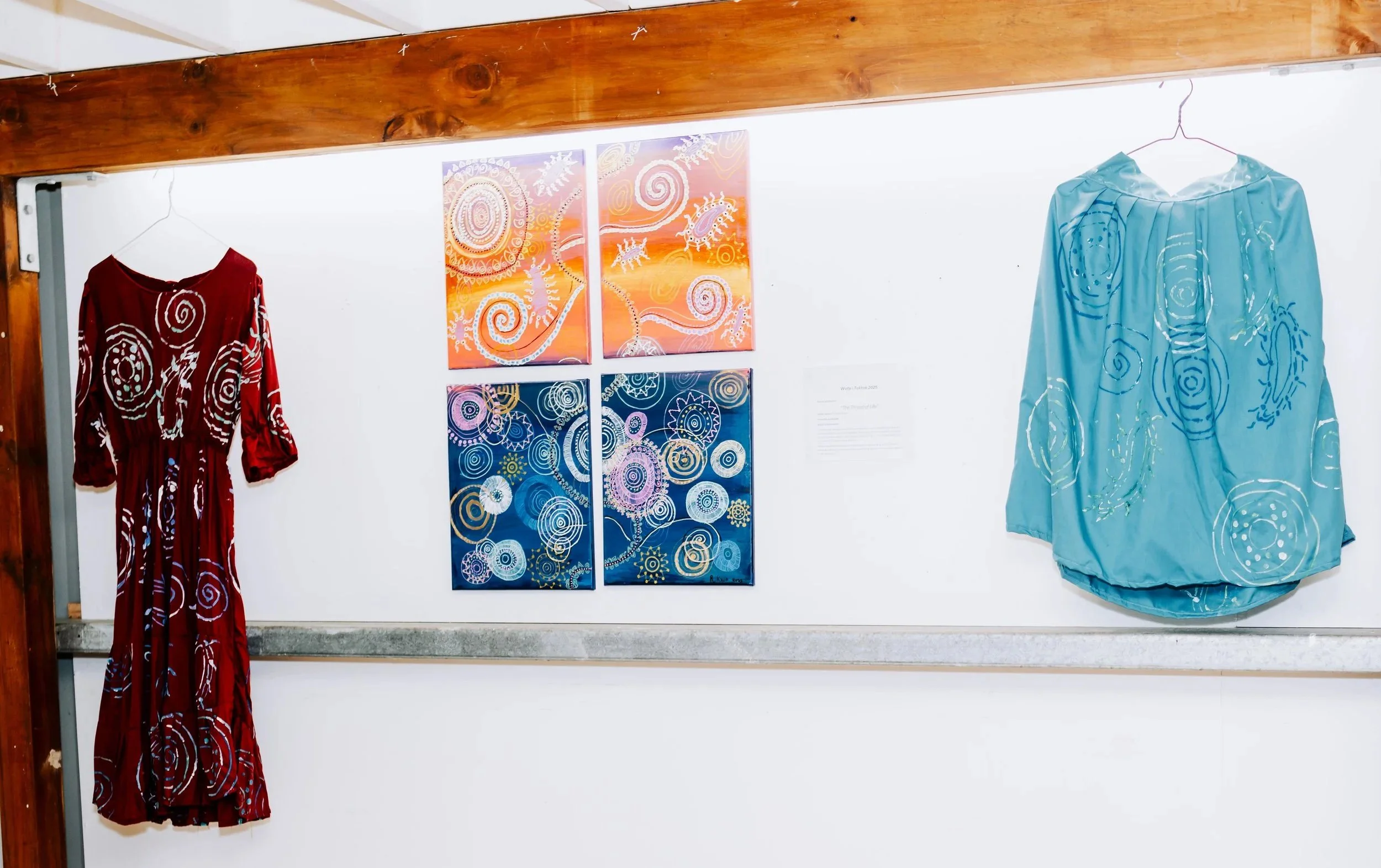Artwork display with four paintings featuring colorful spiral and dotted designs, two dresses hanging on either side with matching abstract patterns, and a white informational plaque on a white wall in a wooden exhibition space.