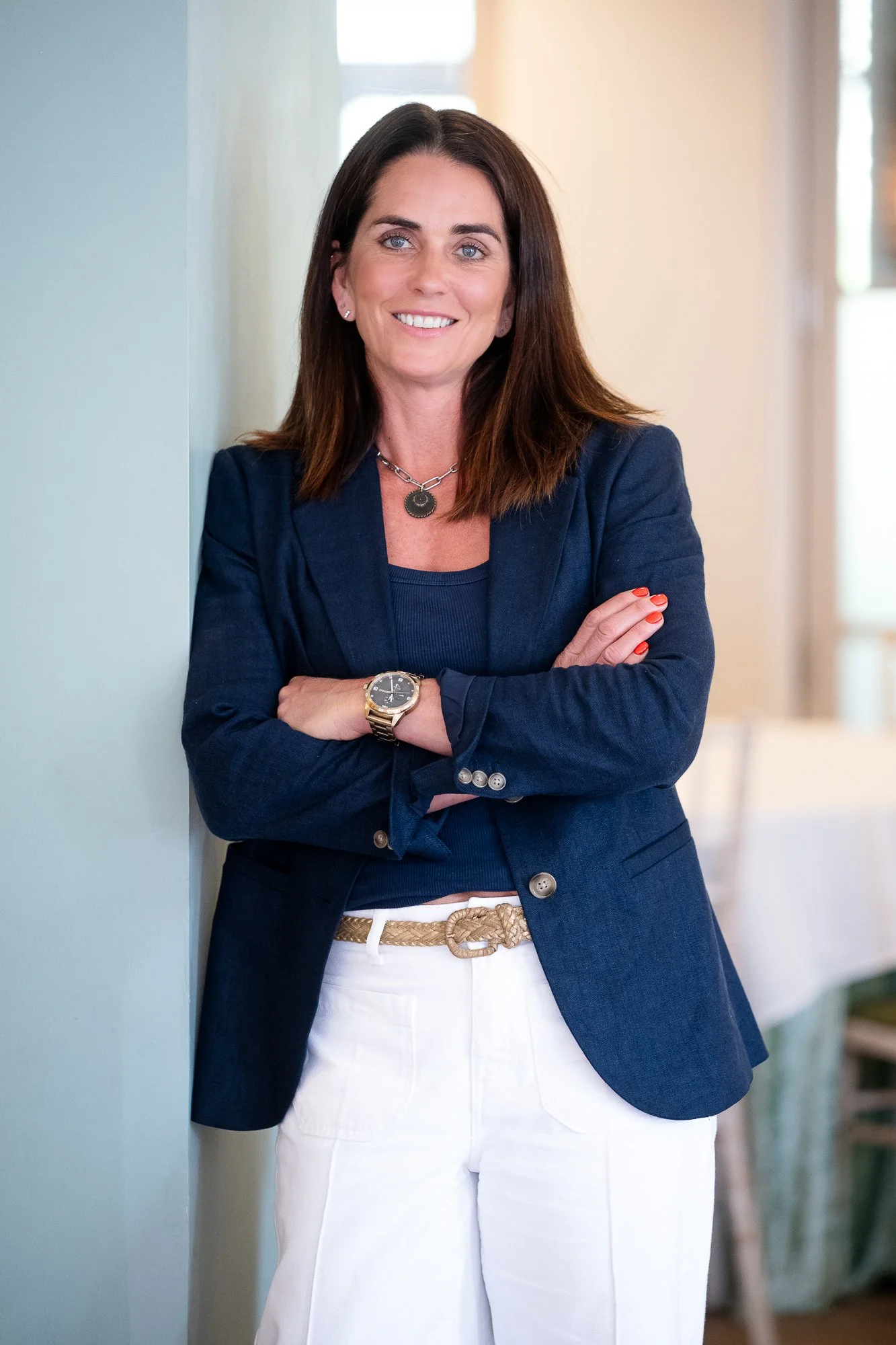 A woman with dark brown hair and blue eyes, wearing a navy blazer, navy top, white pants, and gold accessories, leaning against a wall with arms crossed, smiling.
