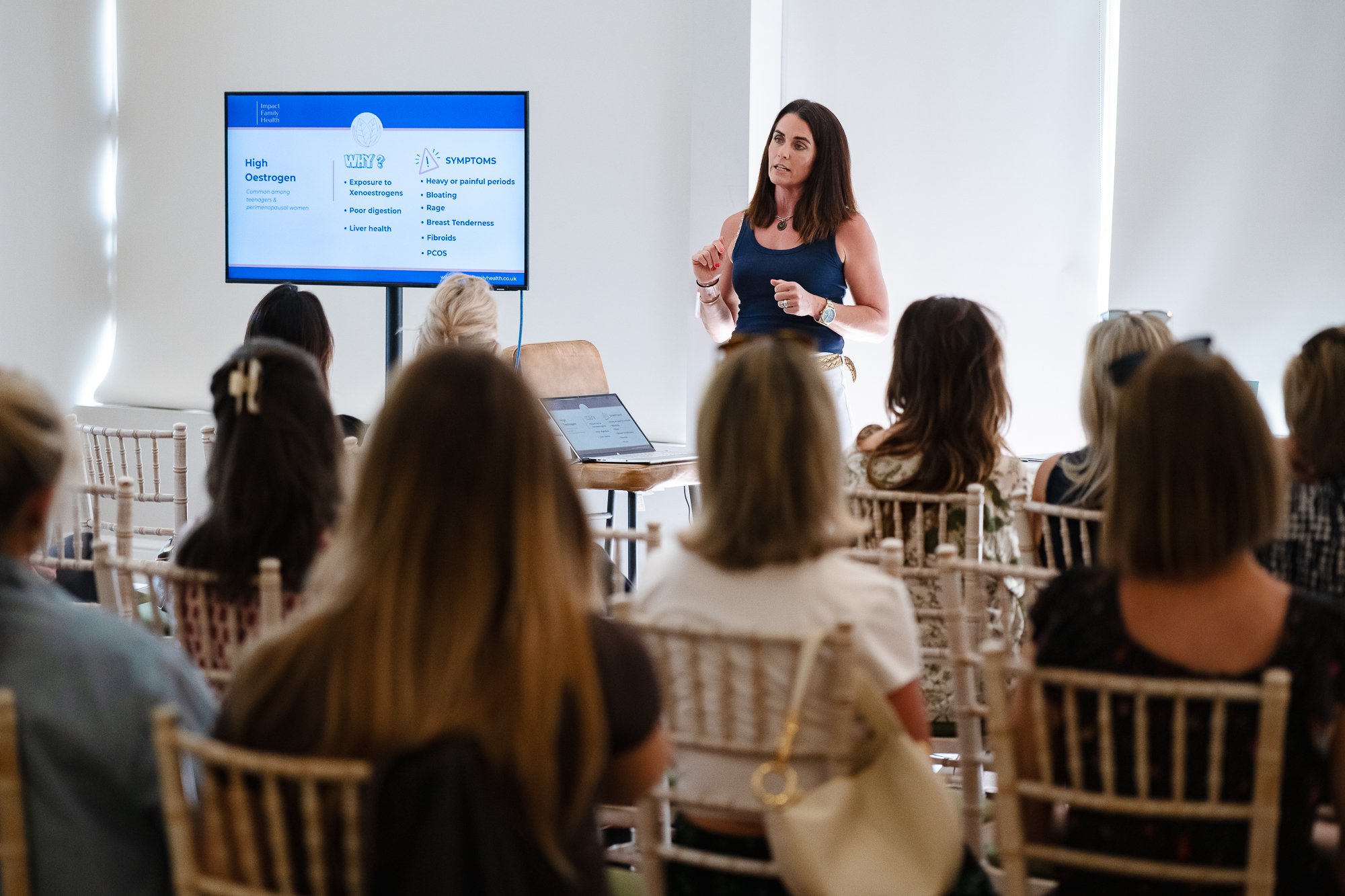 A woman gives a presentation to a group of women in a meeting room. The presentation slide on the screen discusses high estrogen levels and symptoms.