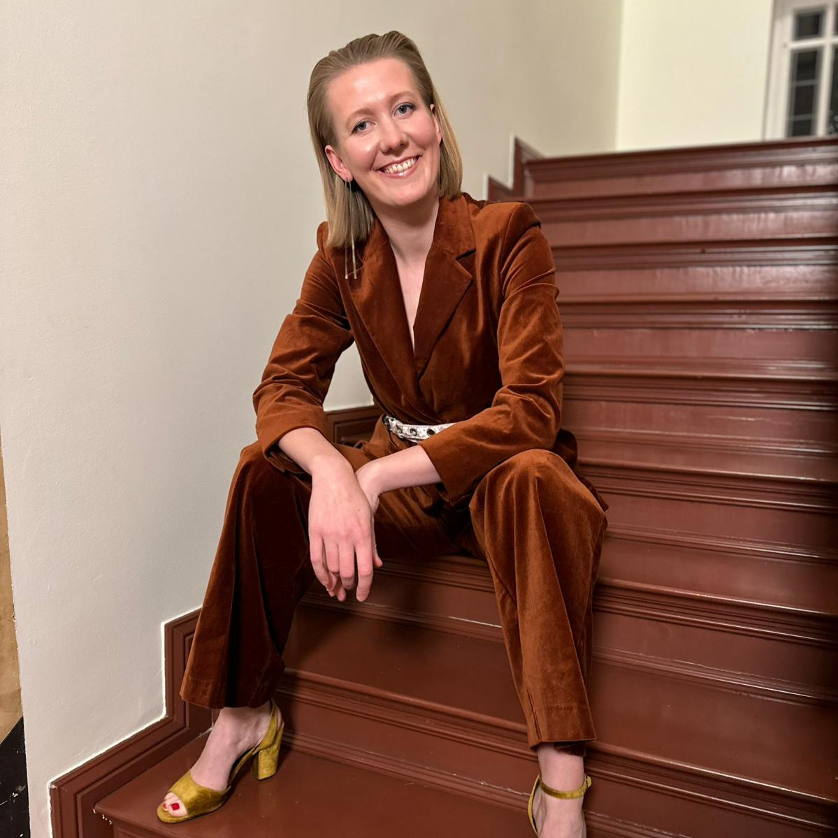 Eine lachende Frau in braunem Anzug sitzt auf einer Holztreppe in einem Innenraum.