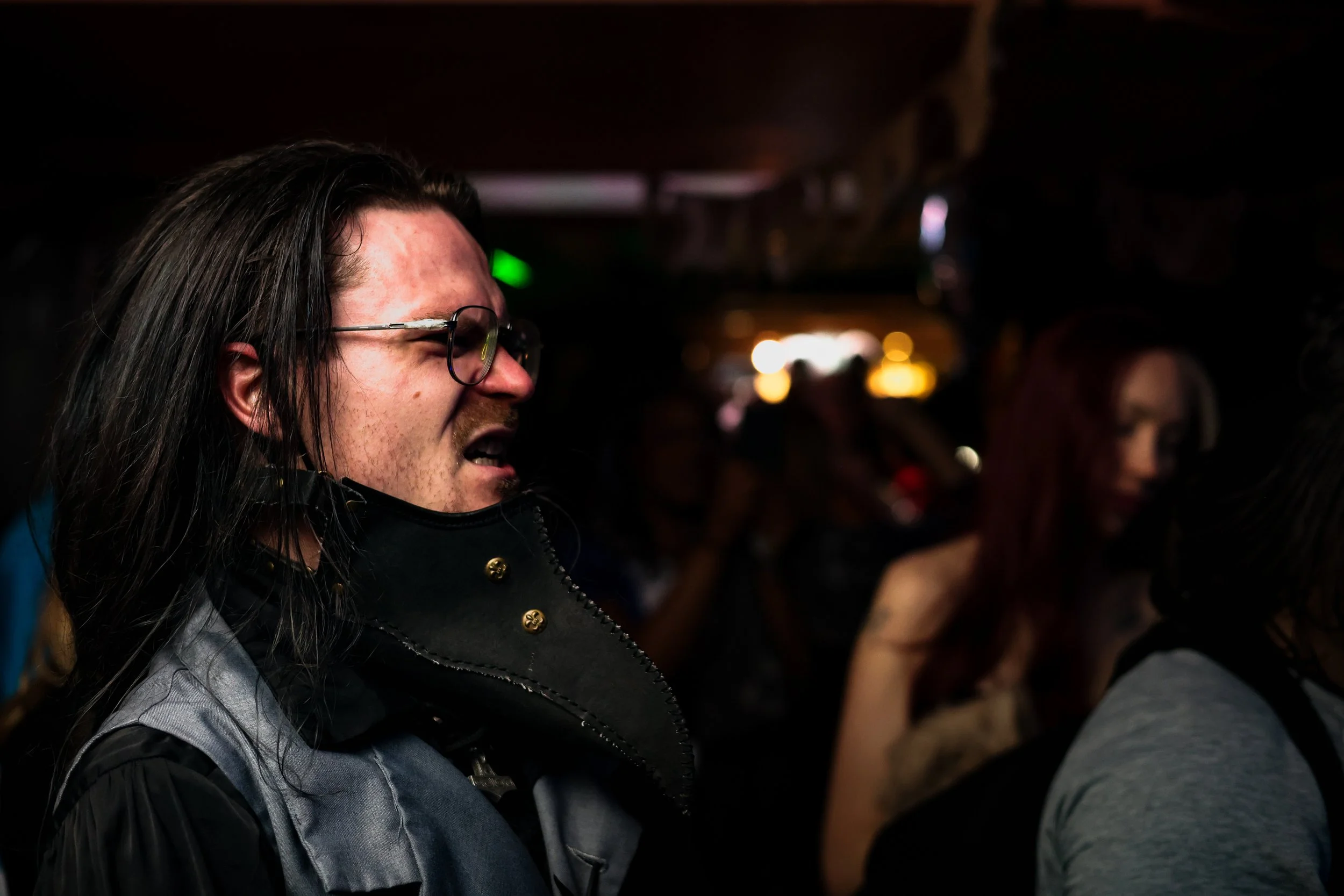 A man with long dark hair, glasses, and a face mask hanging from his ear, appears to be singing or shouting at a crowded event in a dimly lit space.