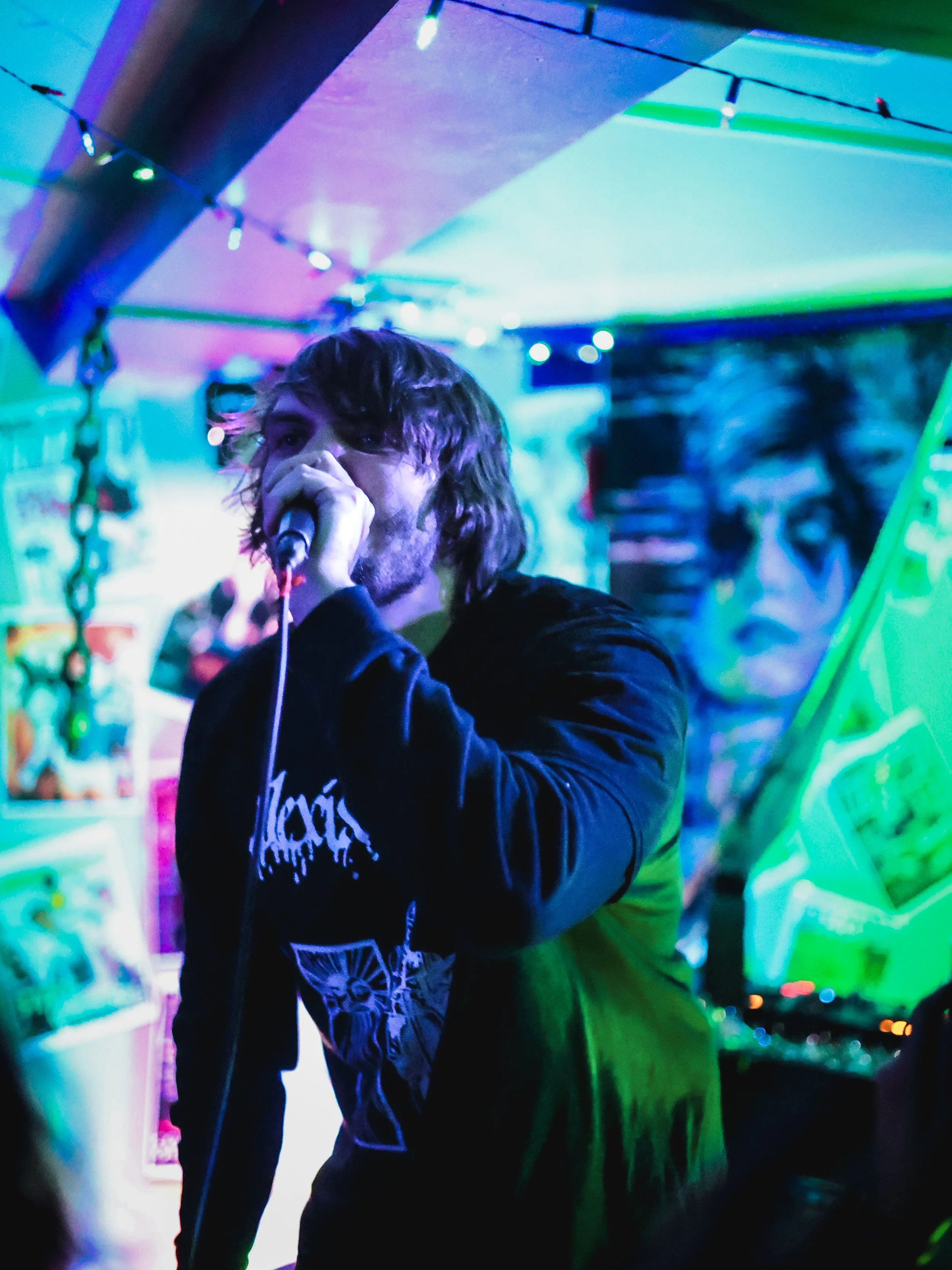 A man with long hair and a beard is singing into a microphone at a music event with colorful, neon lighting and posters in the background.