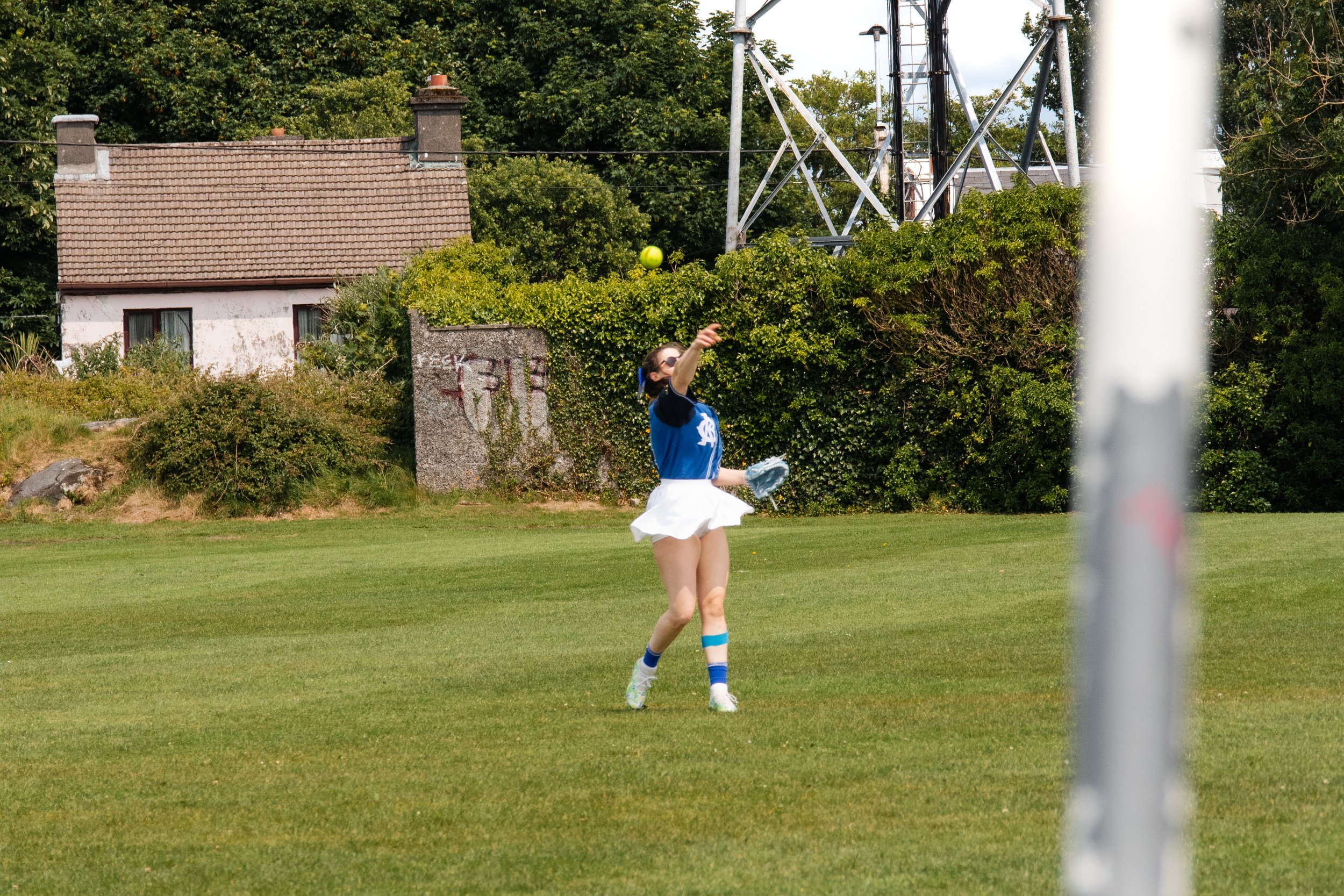 A woman in a blue sports jersey, white skirt, and knee-high socks playing softball outdoors on a grassy field, preparing to throw a yellow softball.