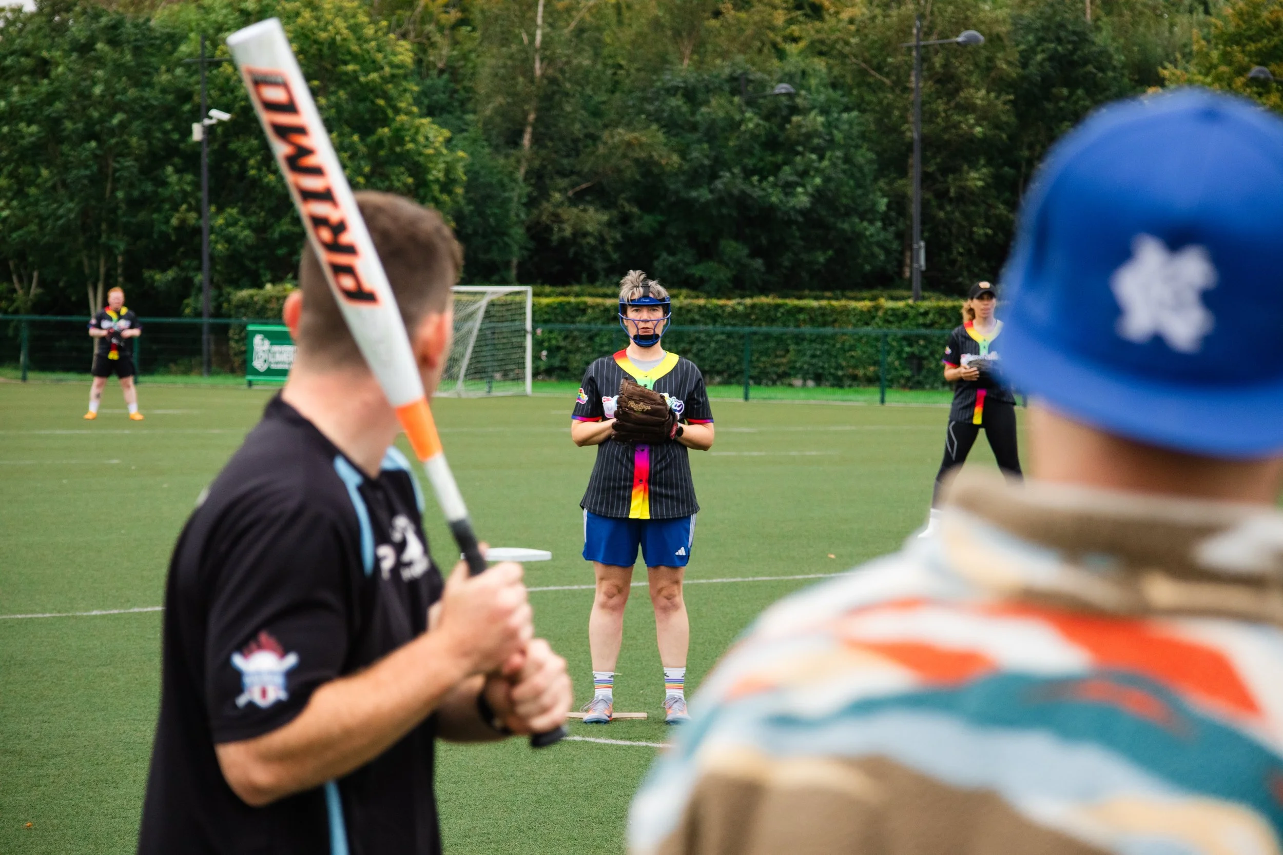 People on a sports field, one person in the foreground holding a baseball bat, one person in the center wearing a helmet and gloves, two additional people in the background, all in sports attire, surrounded by trees.