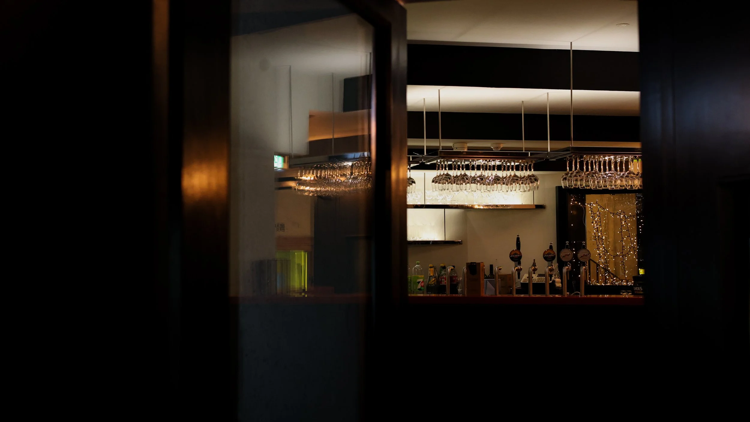 A dimly lit bar area viewed through a doorway, with hanging wine glasses, liquor bottles, and a string of fairy lights in the background.