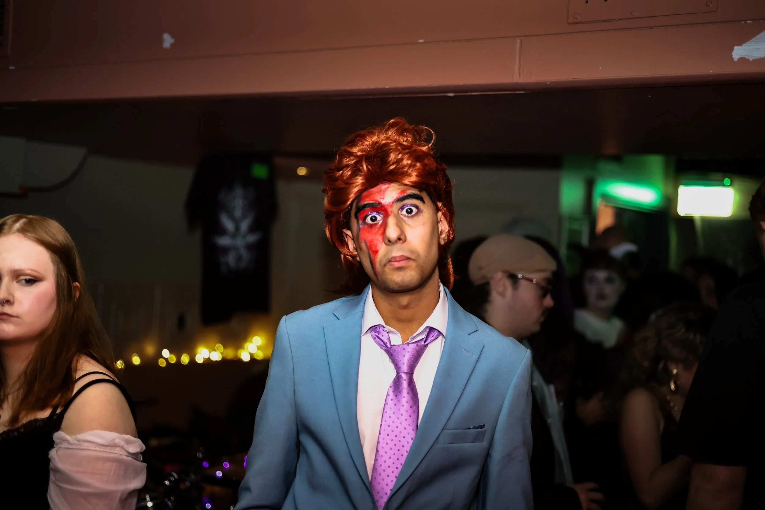 Man in a blue suit with a pink tie, with painted face makeup, at a party or event, surrounded by other people, some with costumes and accessories, in a dimly lit room.