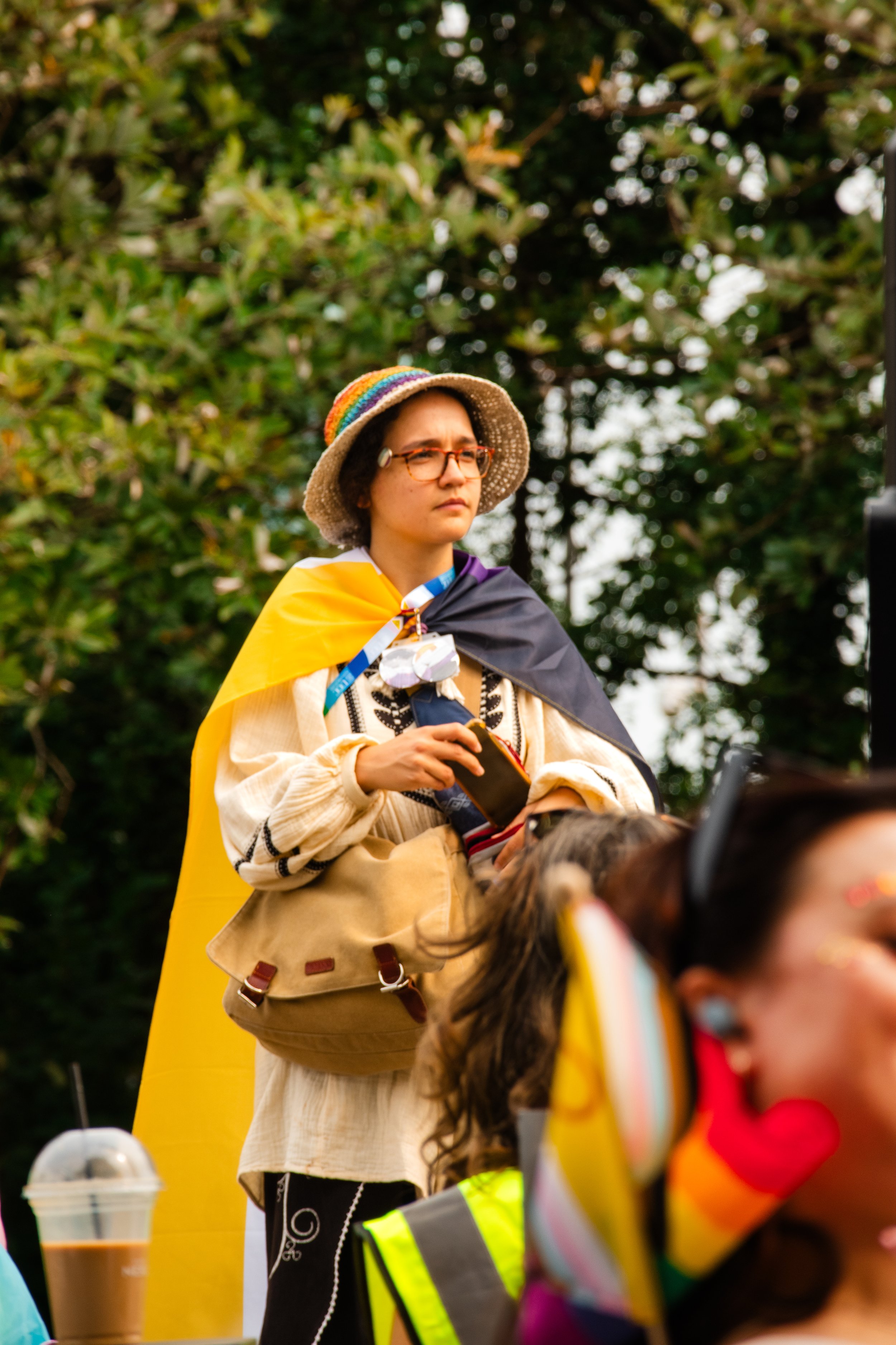 Woman wearing glasses, a rainbow hat, and a cape, holding a phone at an outdoor gathering surrounded by others and greenery.