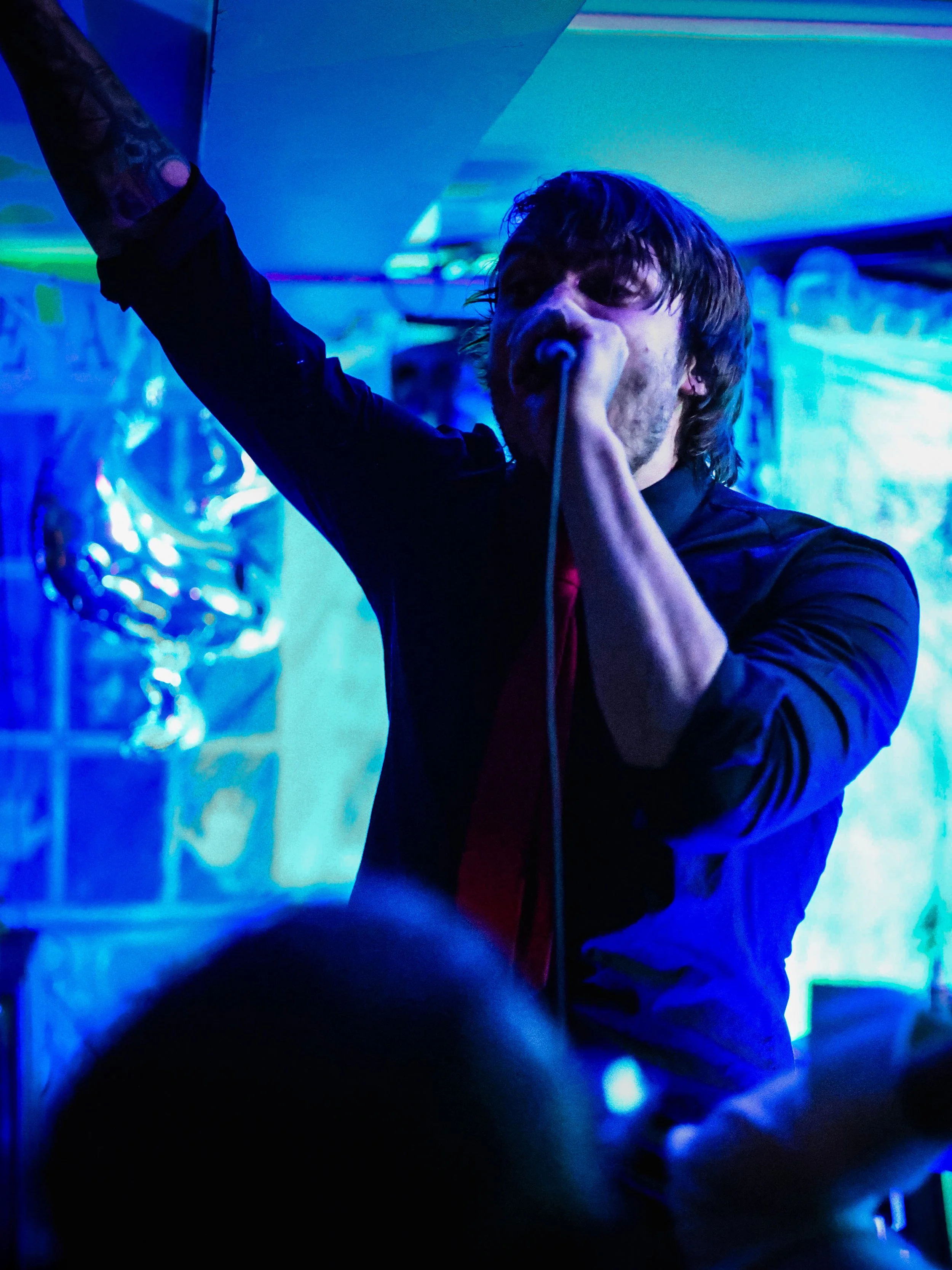 A man with dark, messy hair singing into a microphone at a party or concert, with blue and purple lighting and some balloons in the background.