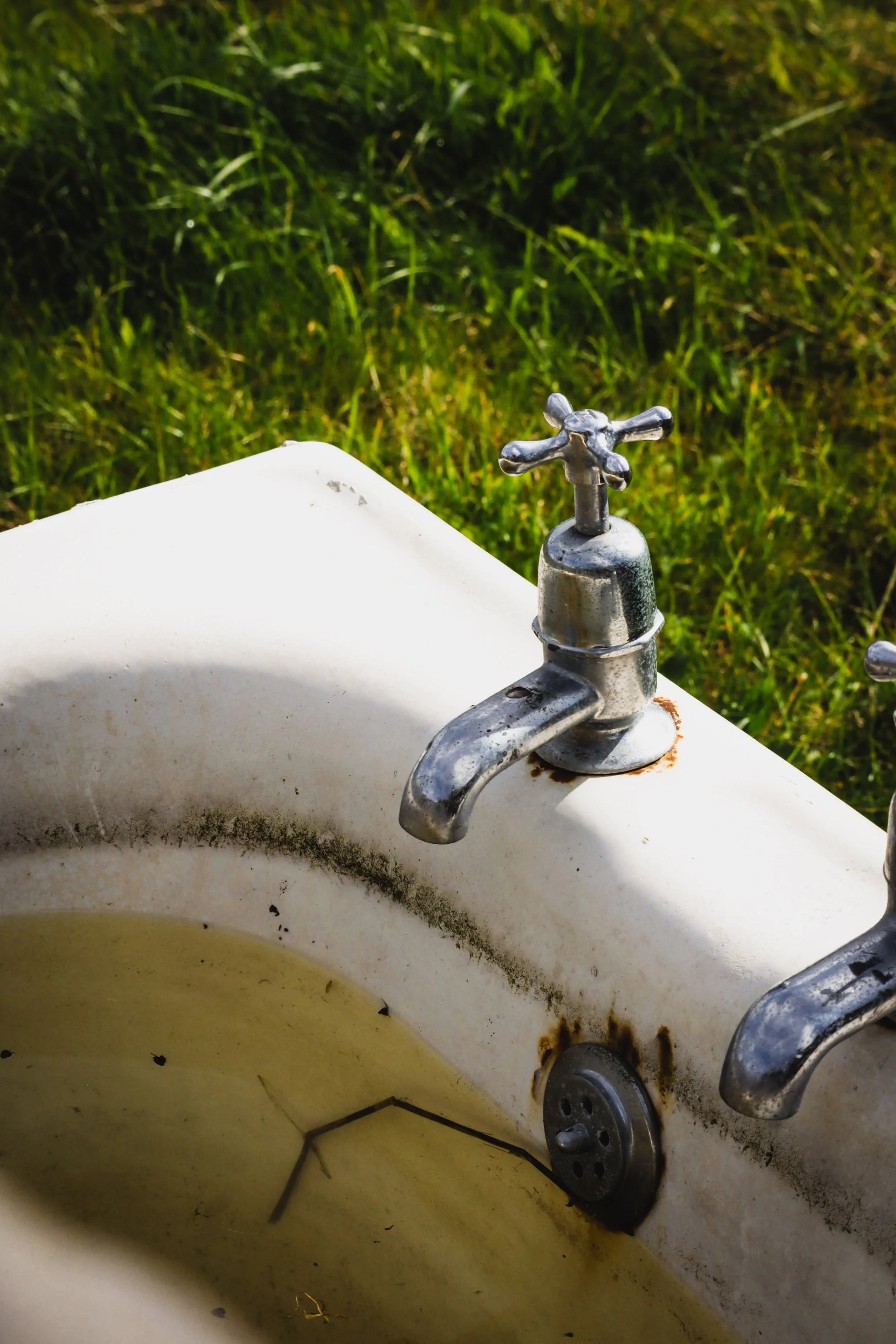 An old outdoor sink with rust and grime, filled with stagnant water, set against a blurred grassy background.