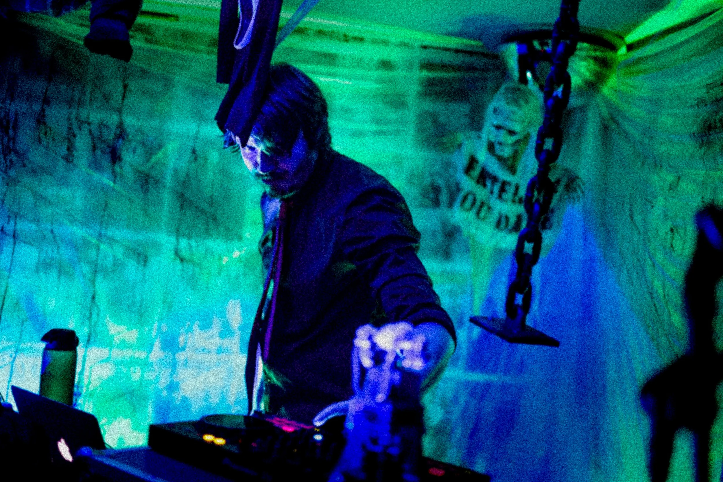 A person with headphones around their neck is DJing in a dimly lit room with colorful neon lighting, a swing hanging from the ceiling, and a 