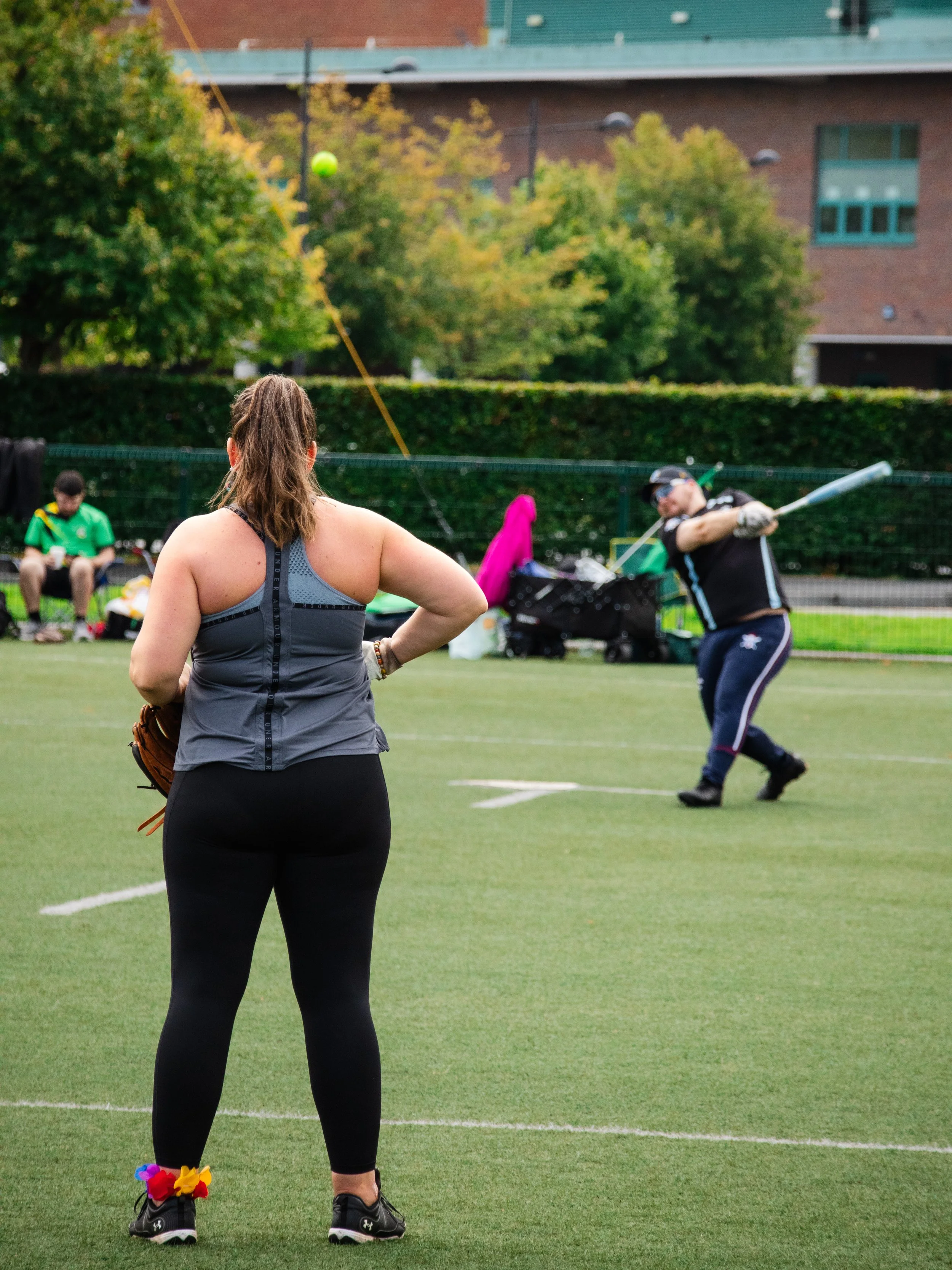 A woman standing on a baseball field watching a man batting, with other people sitting on the sidelines and trees in the background.