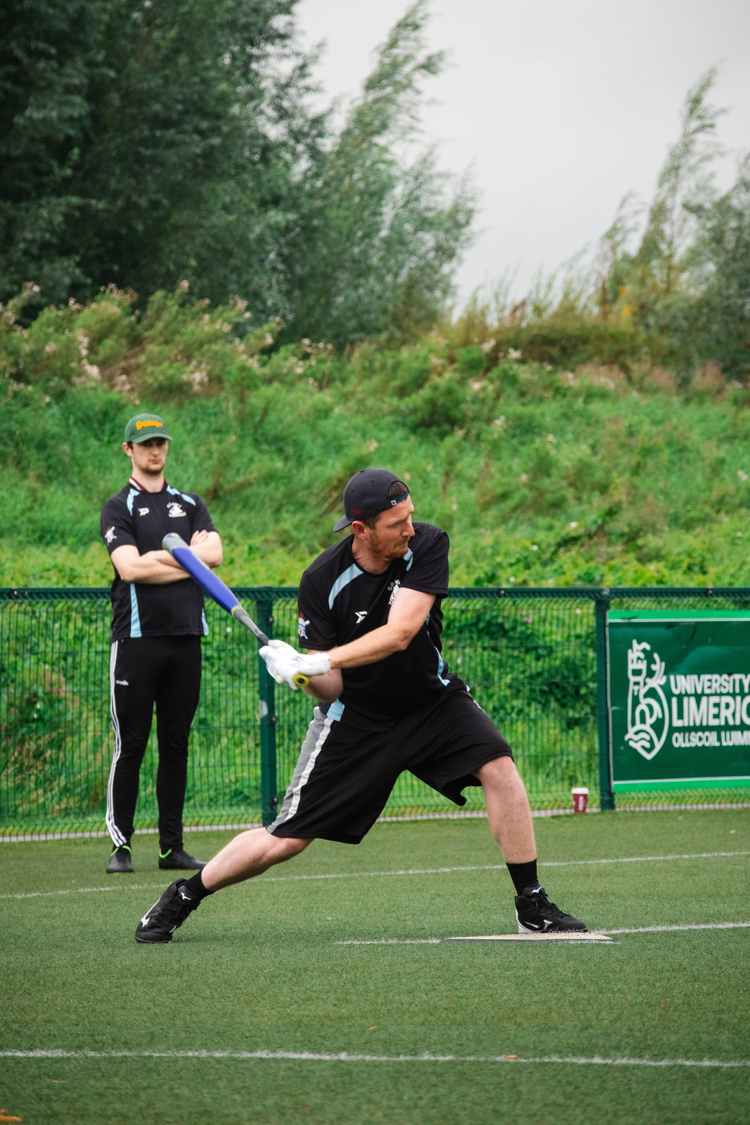 A man in black sportswear swinging a baseball bat on a field, with a woman in similar attire watching behind a fence. There is a sign indicating University of Limerick in the background.