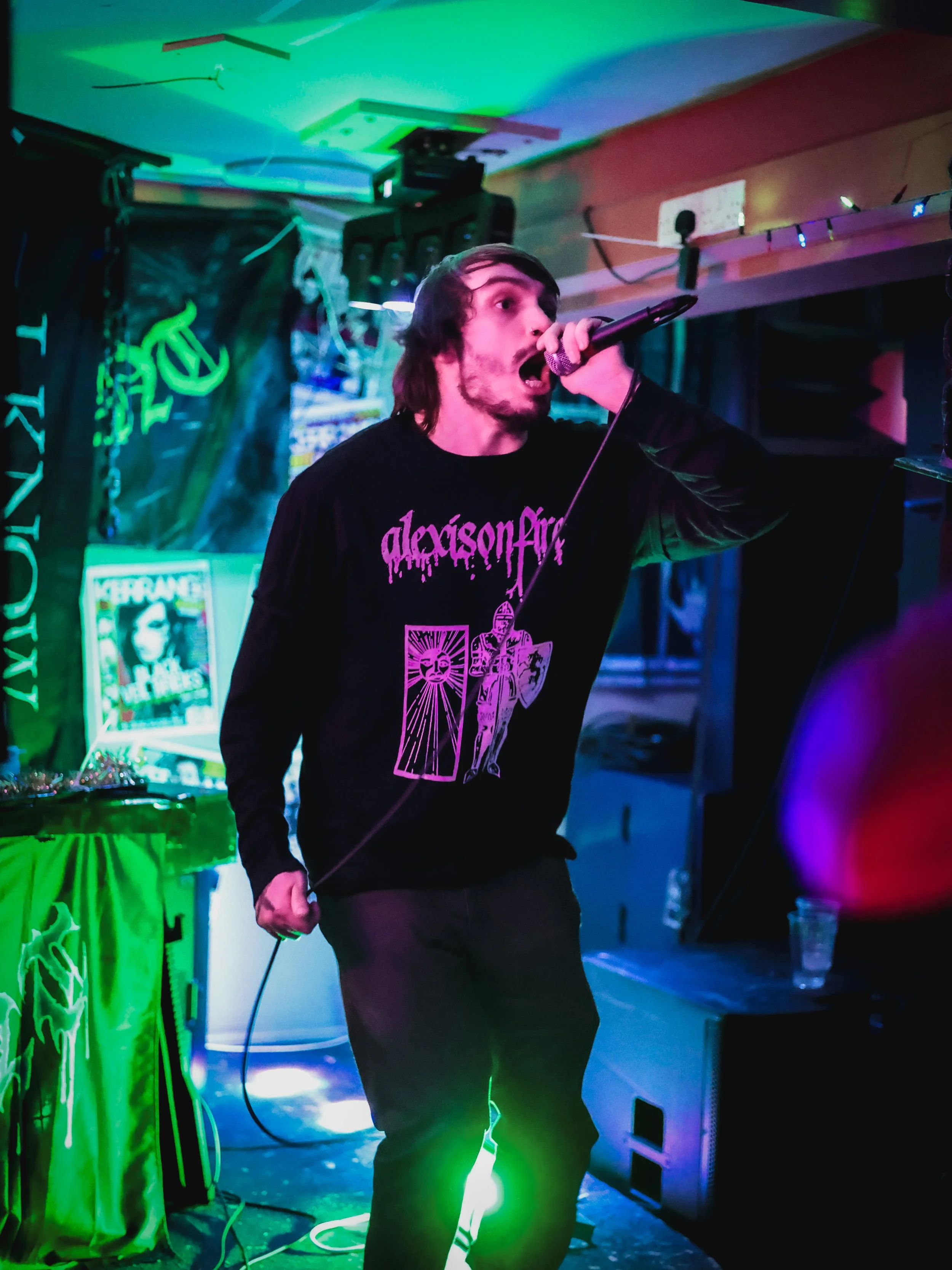 A young man with dark hair and beard singing into a microphone during a live music performance in a dimly lit venue with colorful stage lights, a banner, and posters in the background.