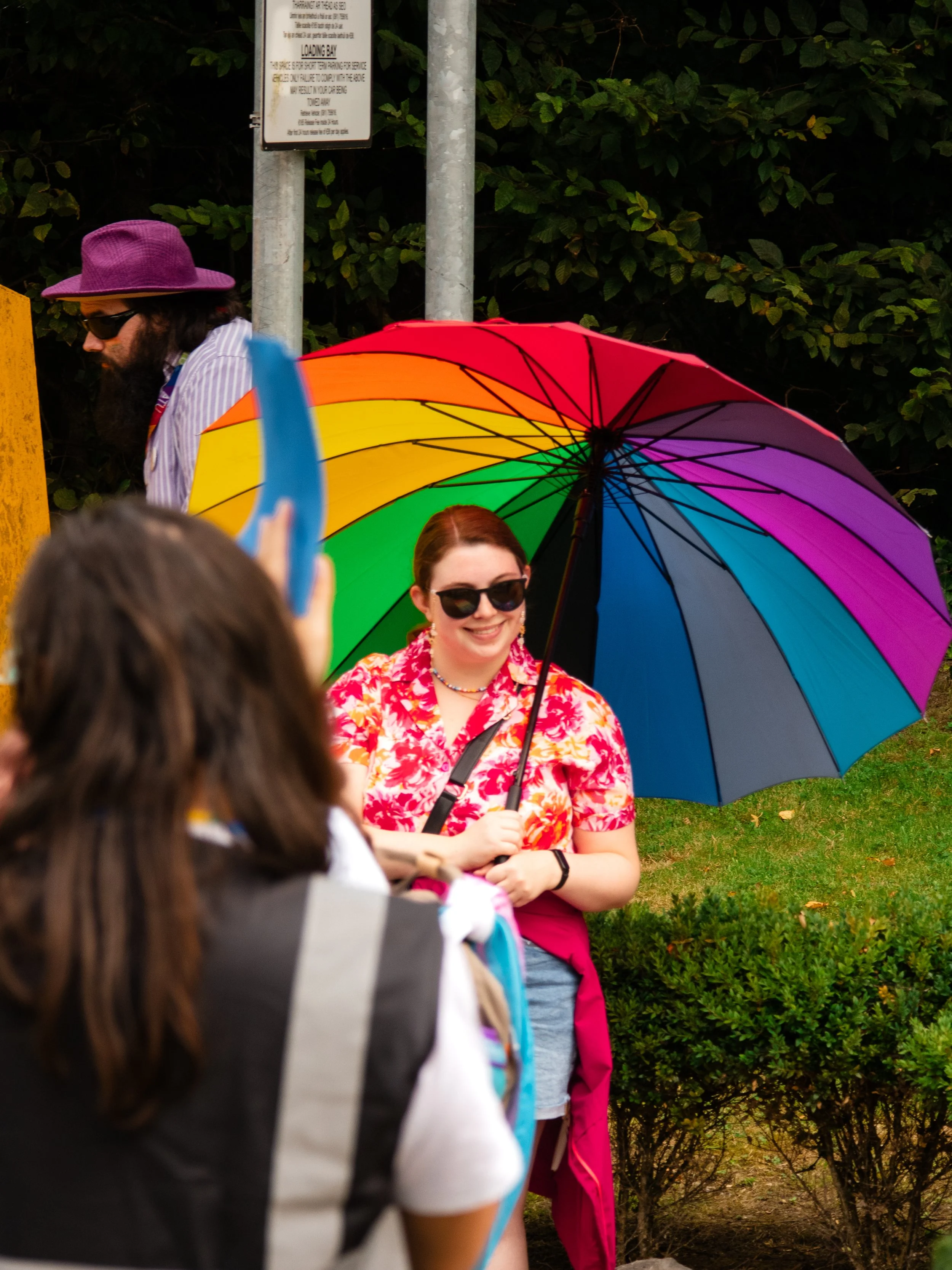 A woman smiling and wearing sunglasses is holding a multicolored rainbow umbrella outdoors, with a person in the foreground partly visible. The background features green bushes and trees.