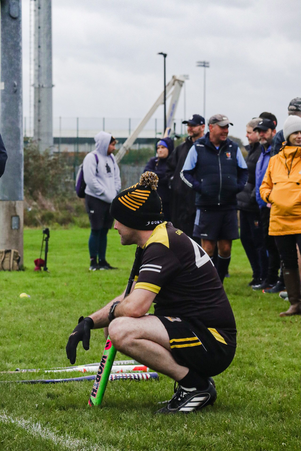 A man squatting on a grassy field holding a colorful cricket bat, wearing a black and yellow sports uniform and a black hat with orange and yellow stripes, while other people dressed in sports and casual clothing stand in the background.