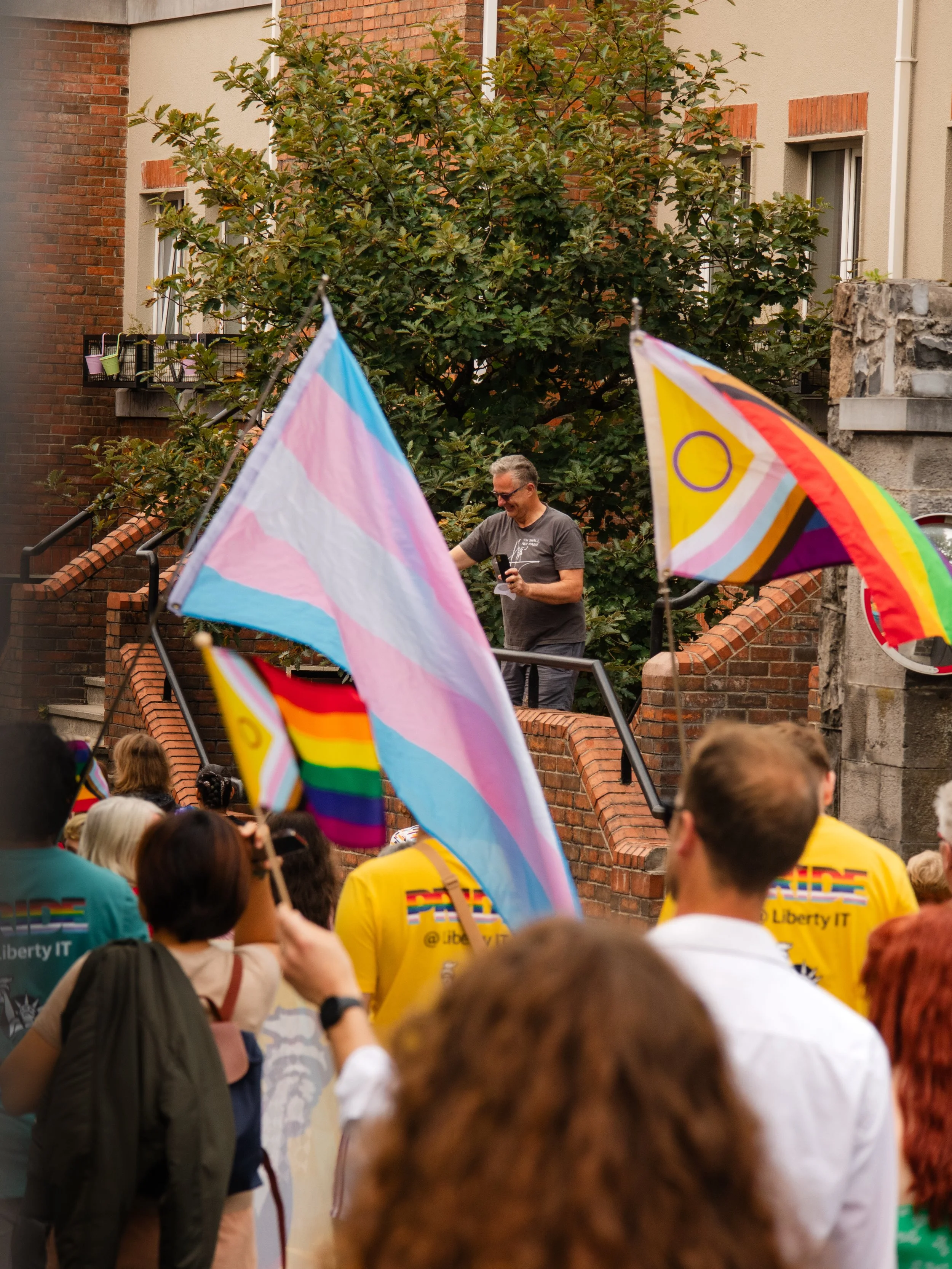 A crowd at a pride parade waving rainbow and transgender flags, with a man on a raised platform addressing the crowd, in front of a brick and concrete building with large trees.