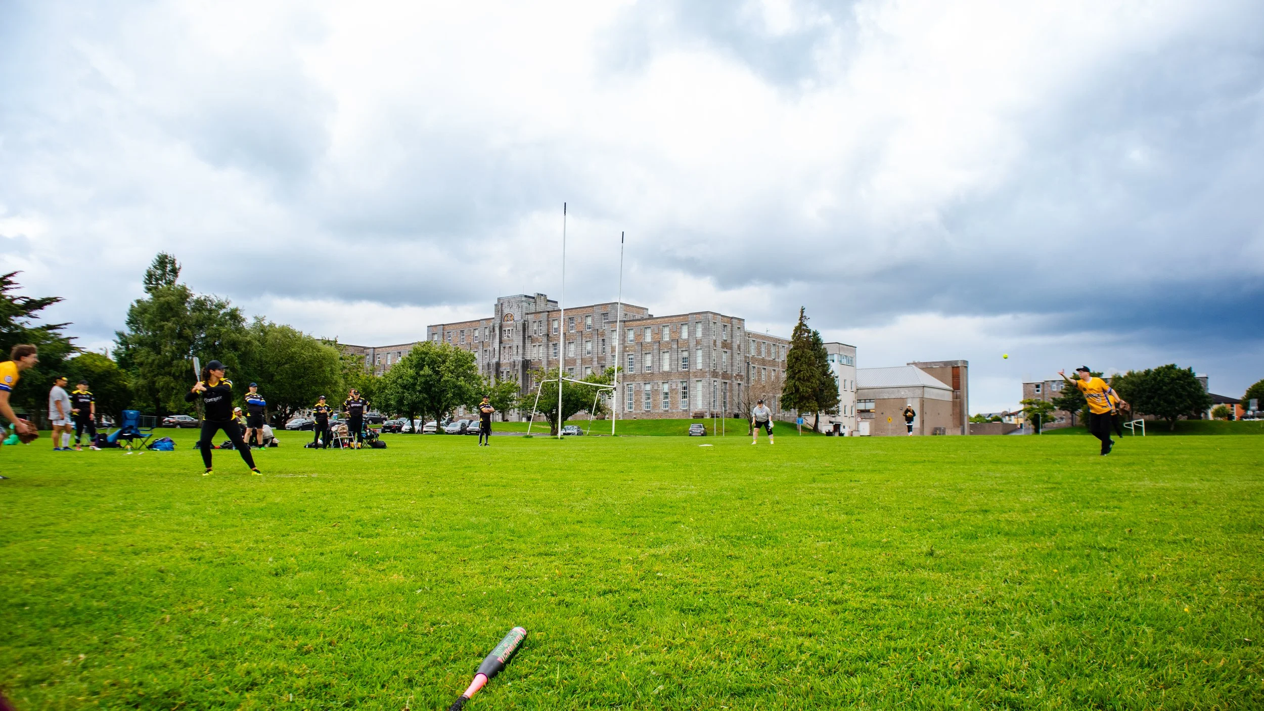 Group of people playing baseball on a grassy field under cloudy sky, with a historic building in the background.