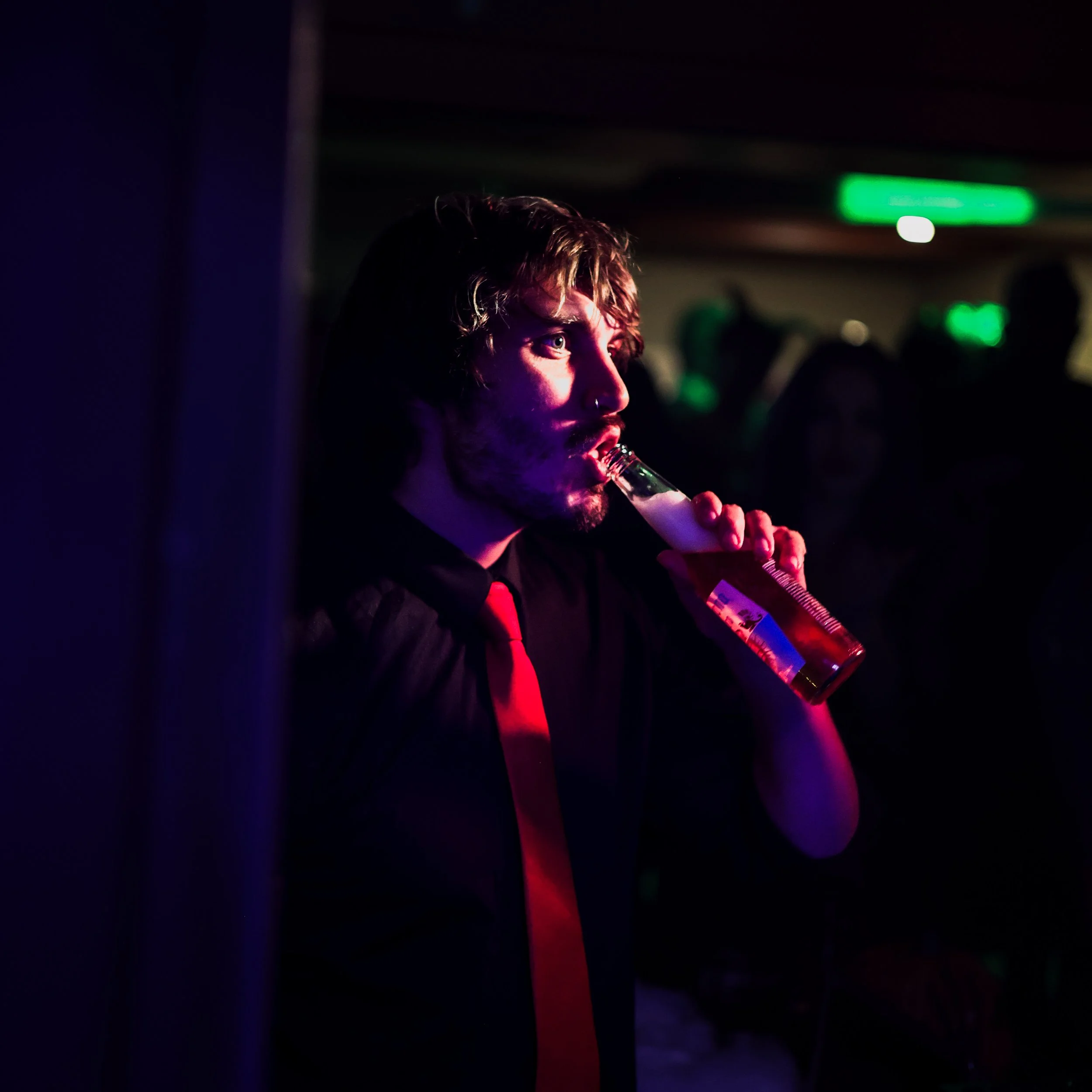 A man with a beard and curly hair in a black shirt and red tie drinking a beer in a dimly lit bar or club with purple and green lighting.
