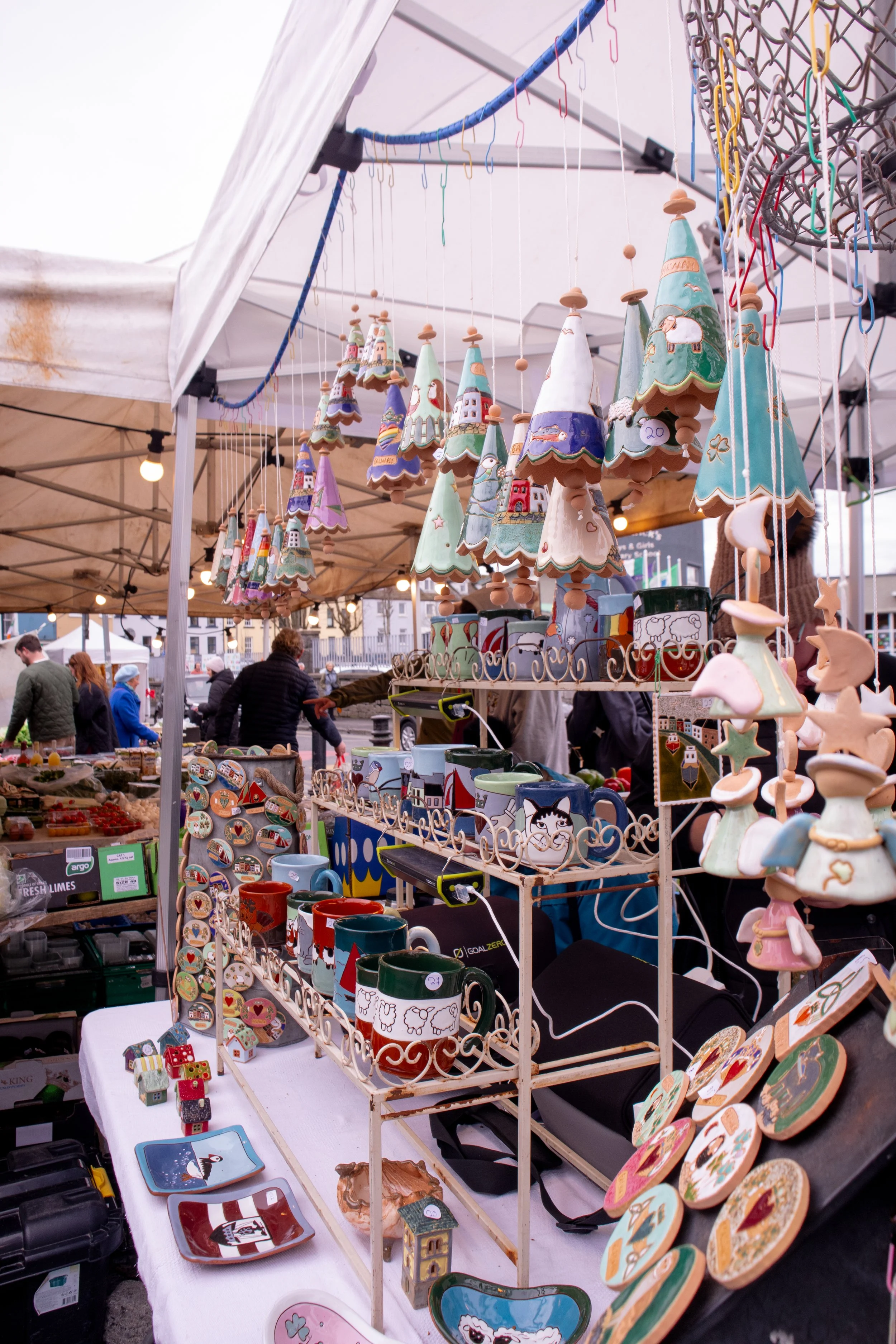A market stall displaying colorful ceramic Christmas tree decorations and holiday-themed mugs with people shopping in the background.