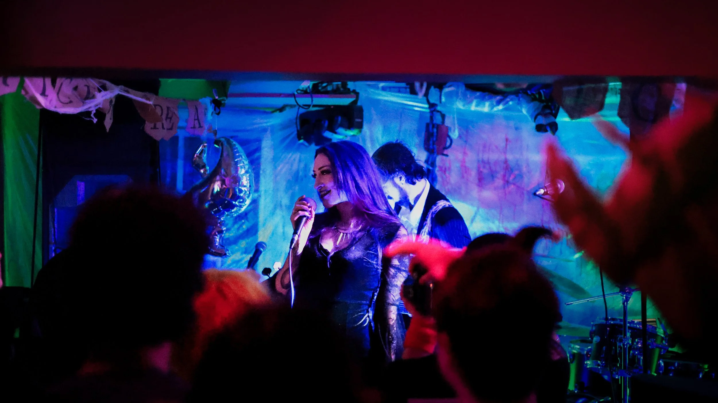 A woman singing into a microphone on stage at a party or celebration, with a man playing drums in the background, colorful decorations, and dim lighting.