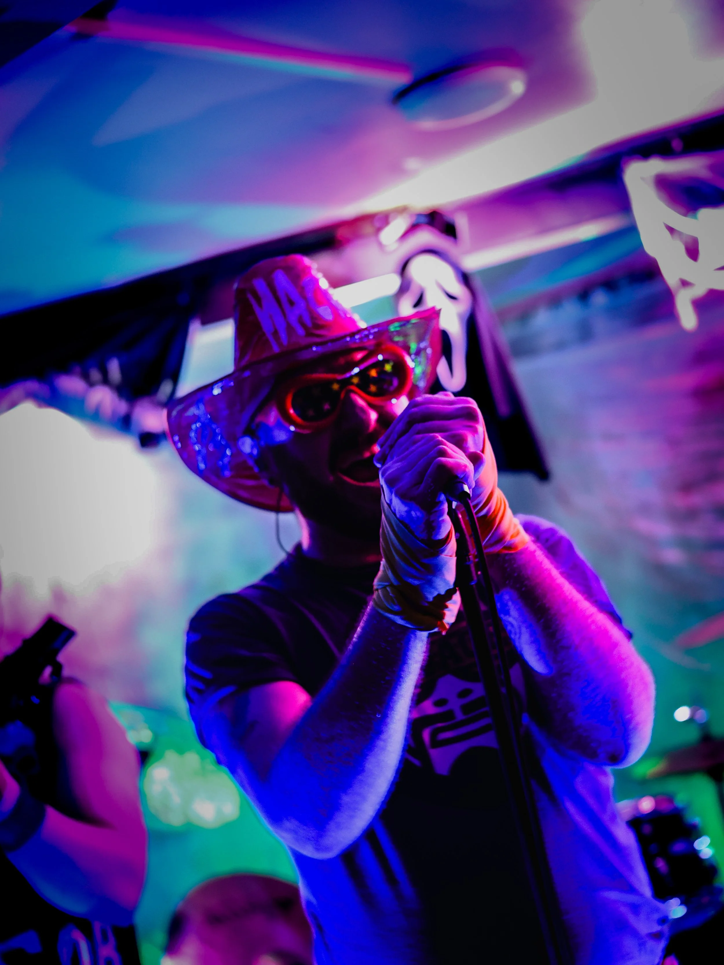 Person wearing a birthday hat and sunglasses singing into a microphone at a lively party or celebration, with colorful lighting and a decorated background.