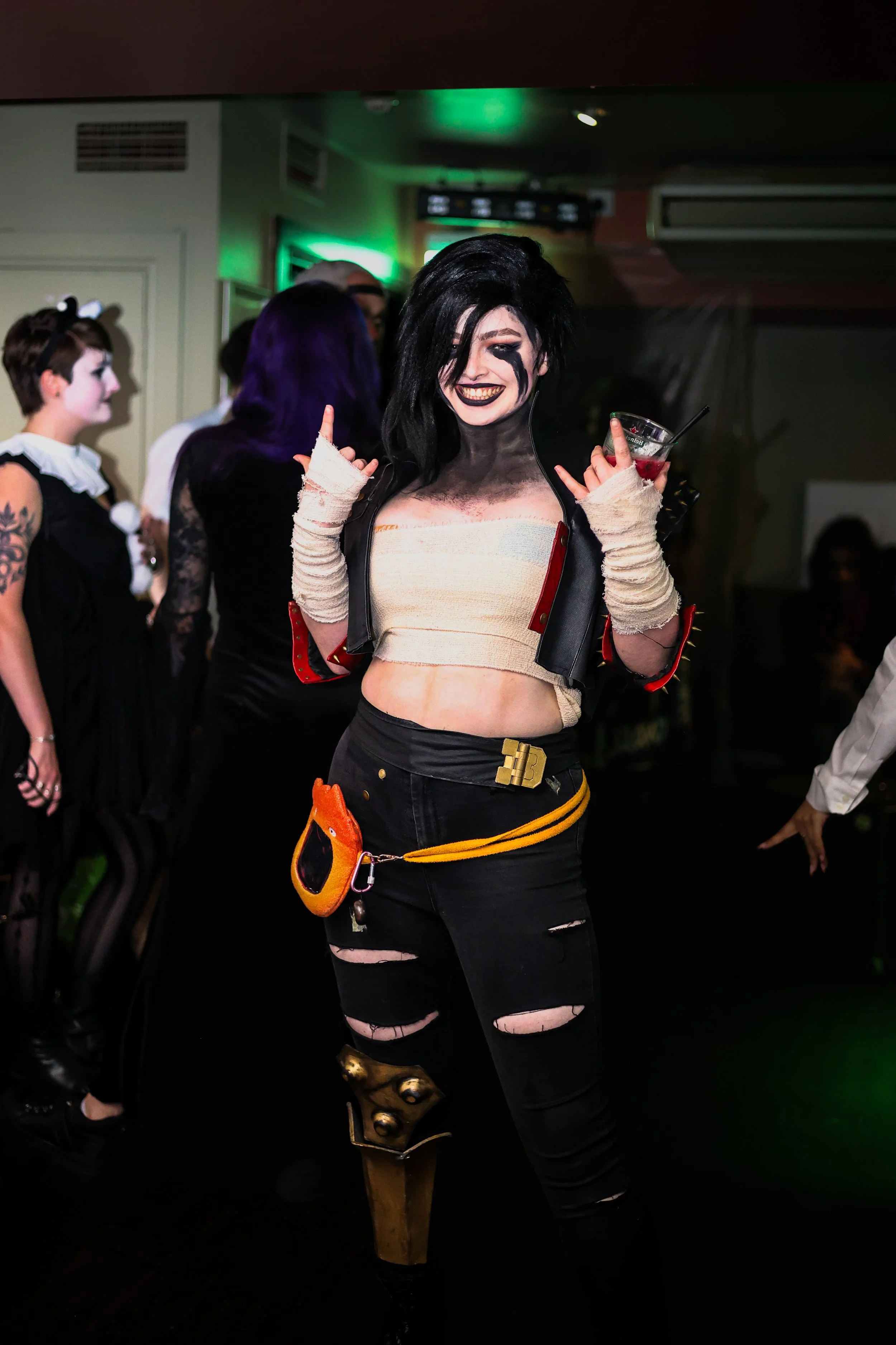 Woman in punk costume with black hair, ripped black pants, and makeup, holding a drink and smiling at a party.