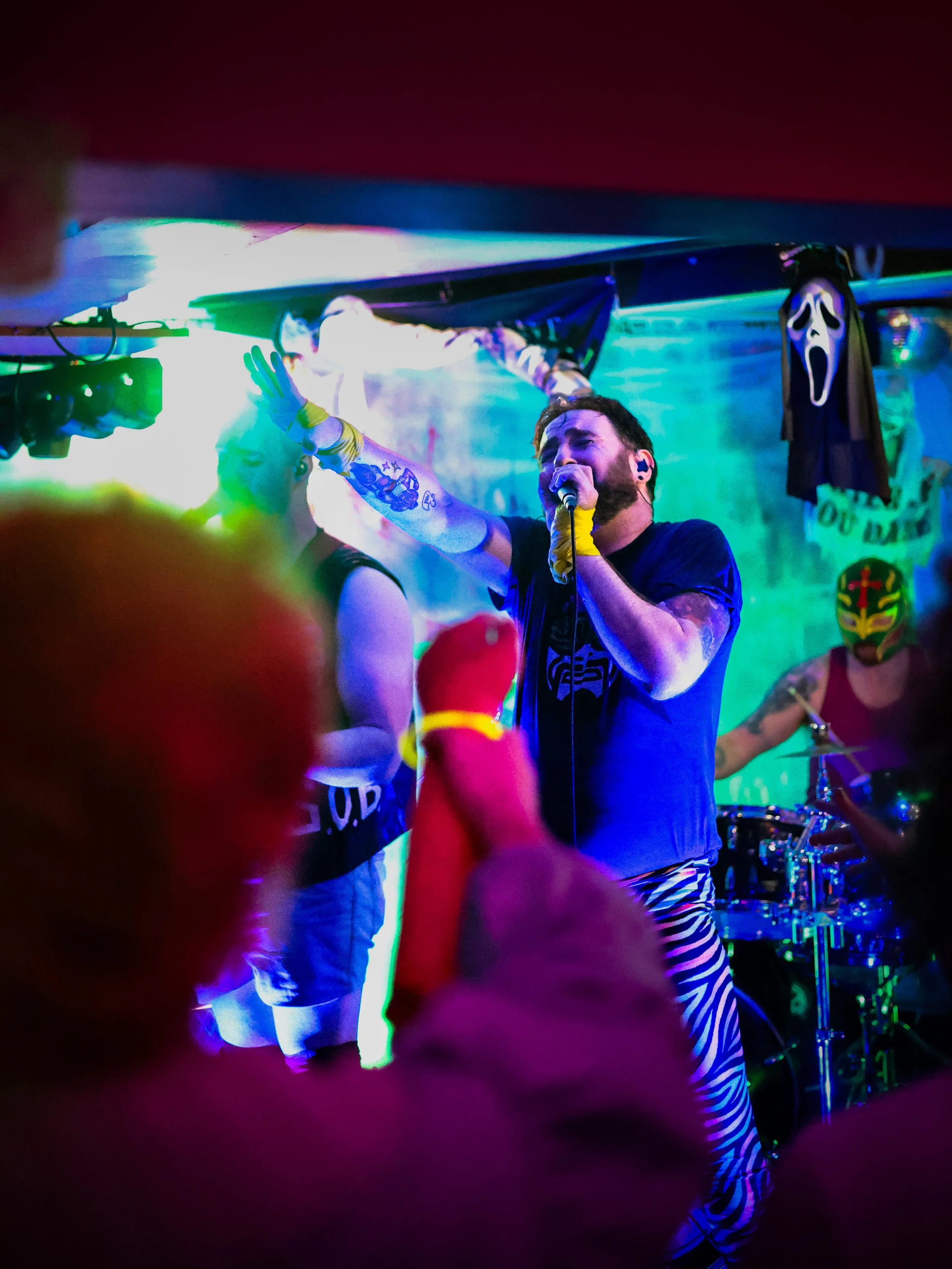 A band performs on stage with colorful lights, including a singer in front with a microphone and striped pants, another performer with a tattooed arm, and a drummer with a masked face in the background; Halloween decorations are visible.