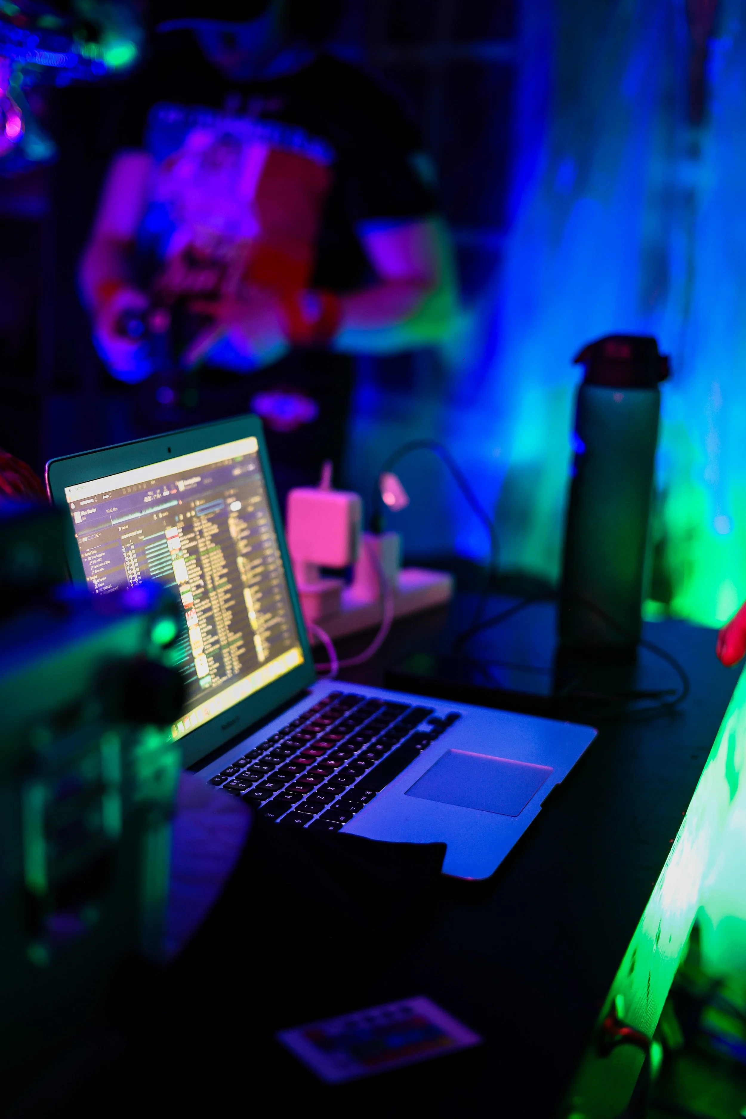 A laptop on a table with music playlist visible on the screen, surrounded by colorful neon lights, drink bottles, and electronic equipment in a dimly lit nightclub or party setting.