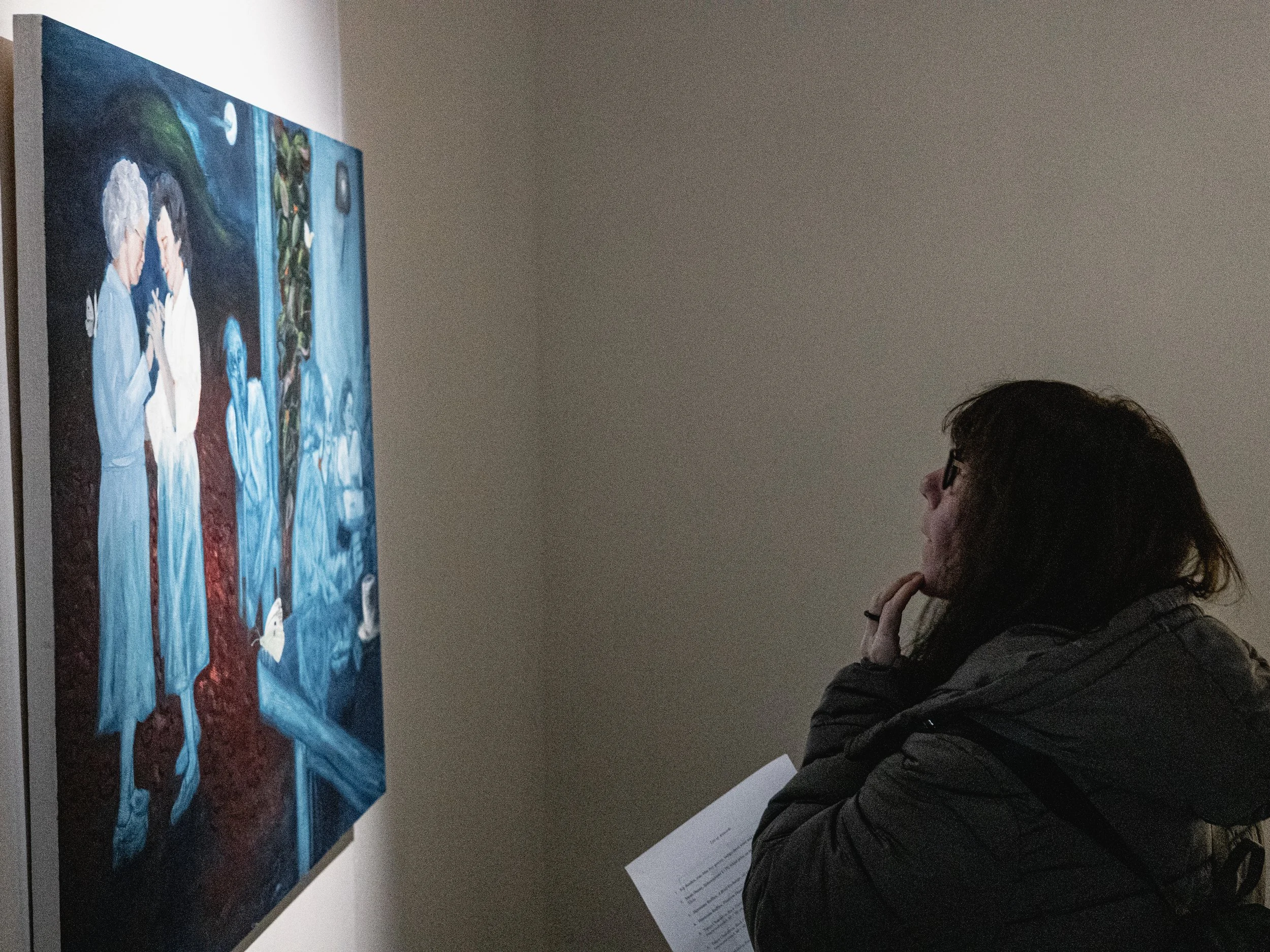 A woman with dark hair, glasses, and a dark jacket is observing a large colorful painting in a gallery. She is holding a paper or catalog in her hand and appears to be thoughtfully studying the artwork.