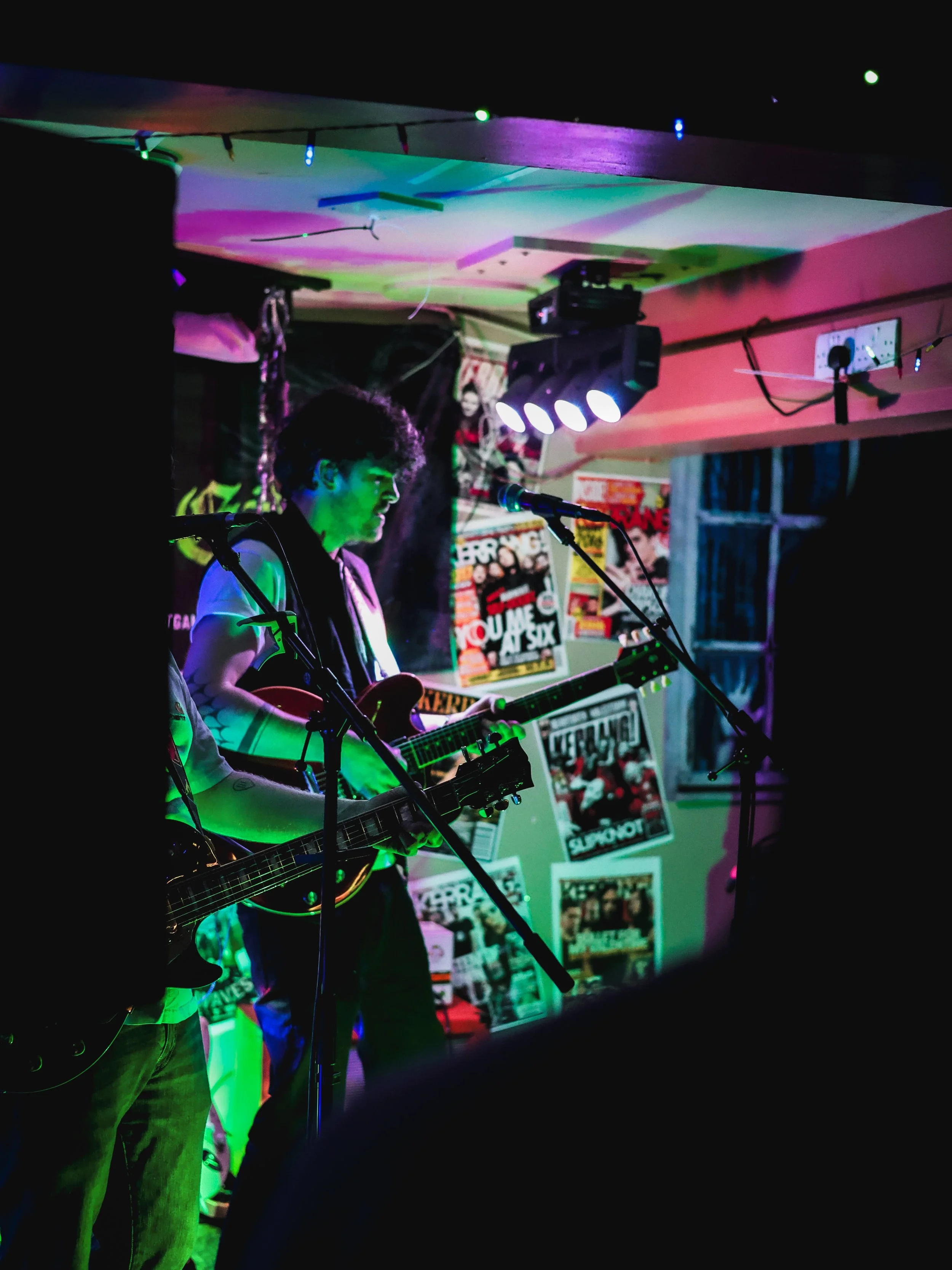 Musicians performing with guitars on a stage decorated with posters under colorful stage lighting.