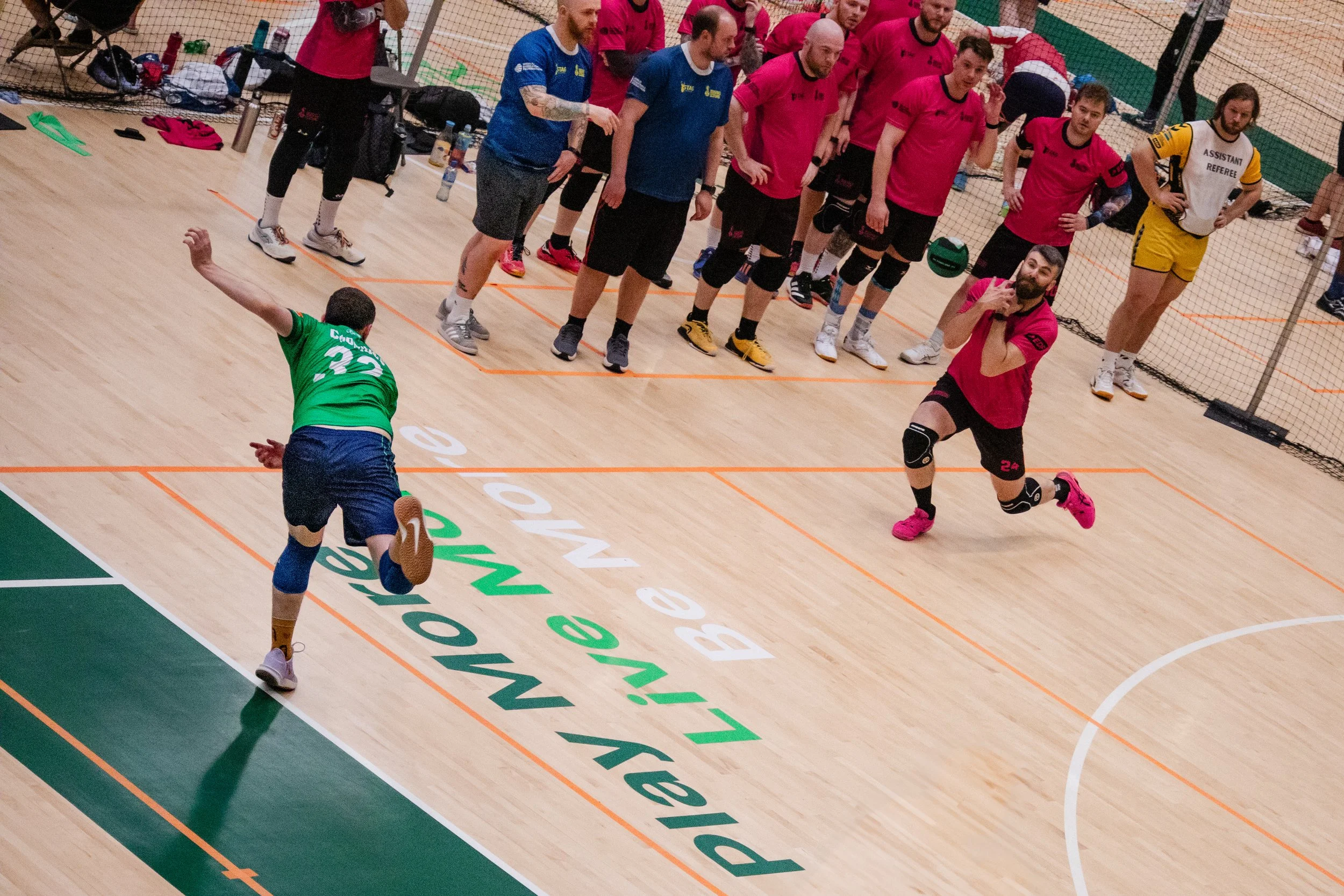 An indoor volleyball match with one player in a green jersey serving the ball while jumping, and another player in a pink jersey, knee brace, pink shoes, and kneeling position ready to receive. Several other players and a referee are visible in the b