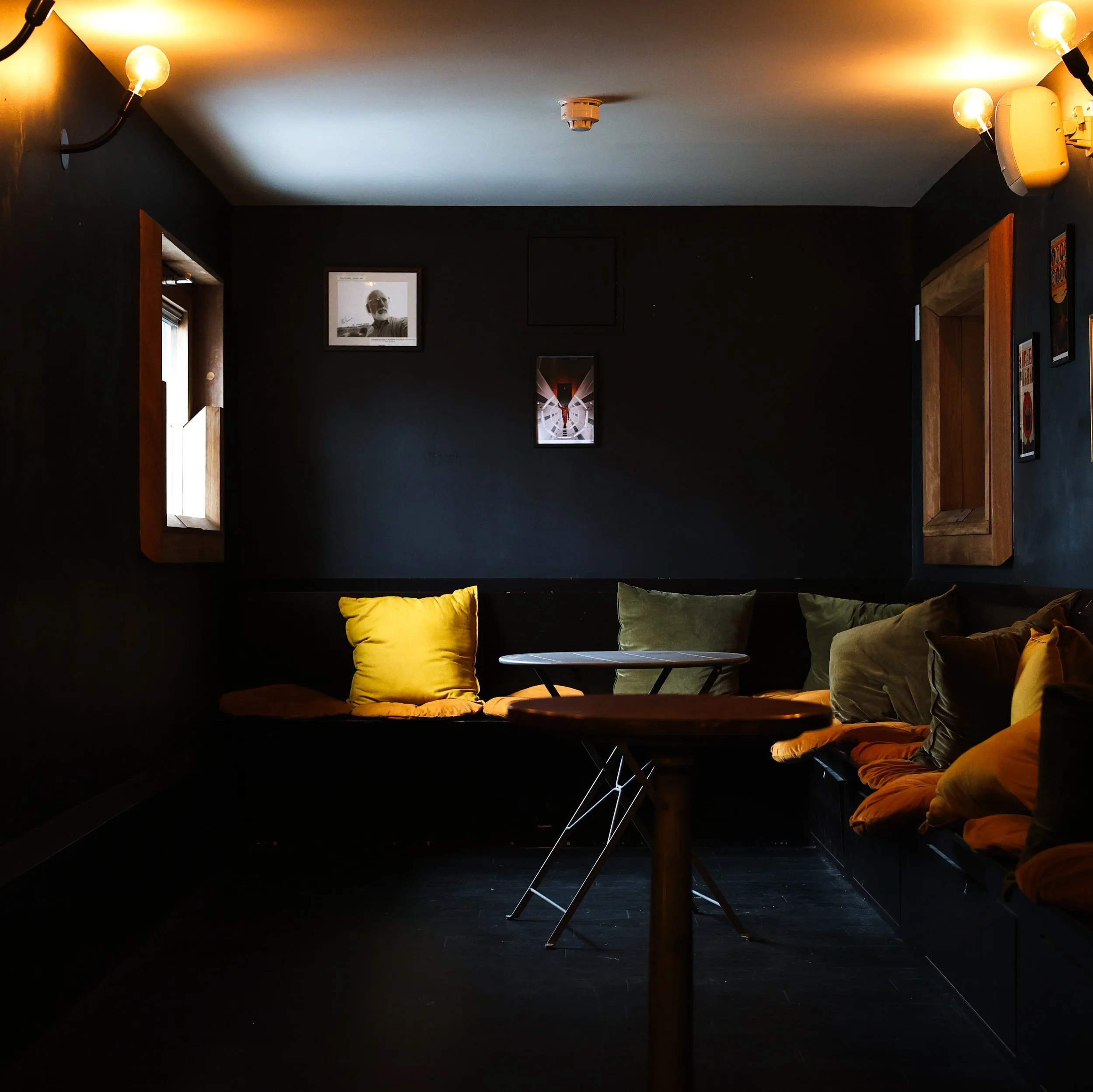 A cozy corner of a dimly lit cafe with black walls decorated with framed pictures. There are yellow and green cushions on a built-in bench and a small round table in the center.