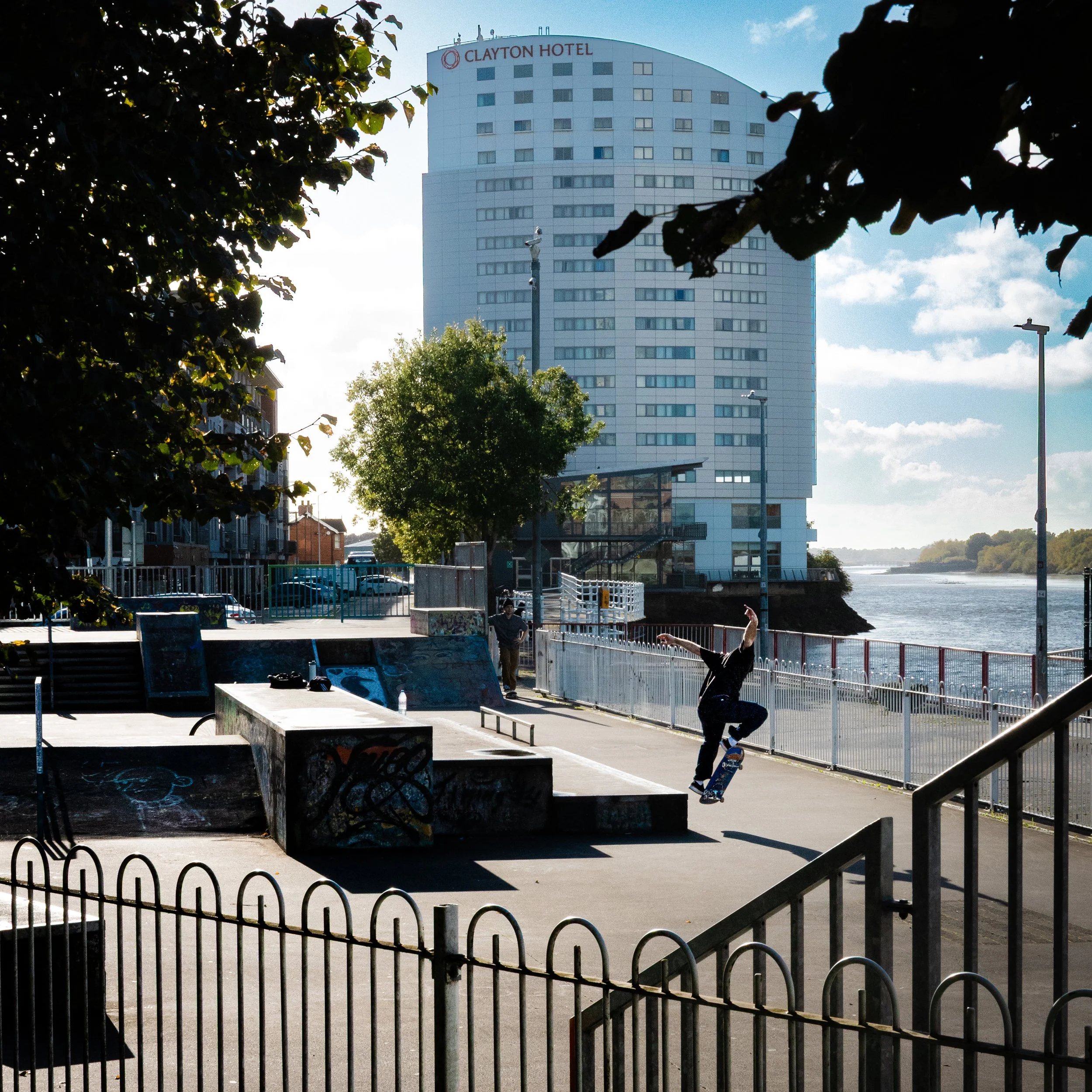 Skateboarder performing a trick at a skate park near a river with a tall hotel building in the background.