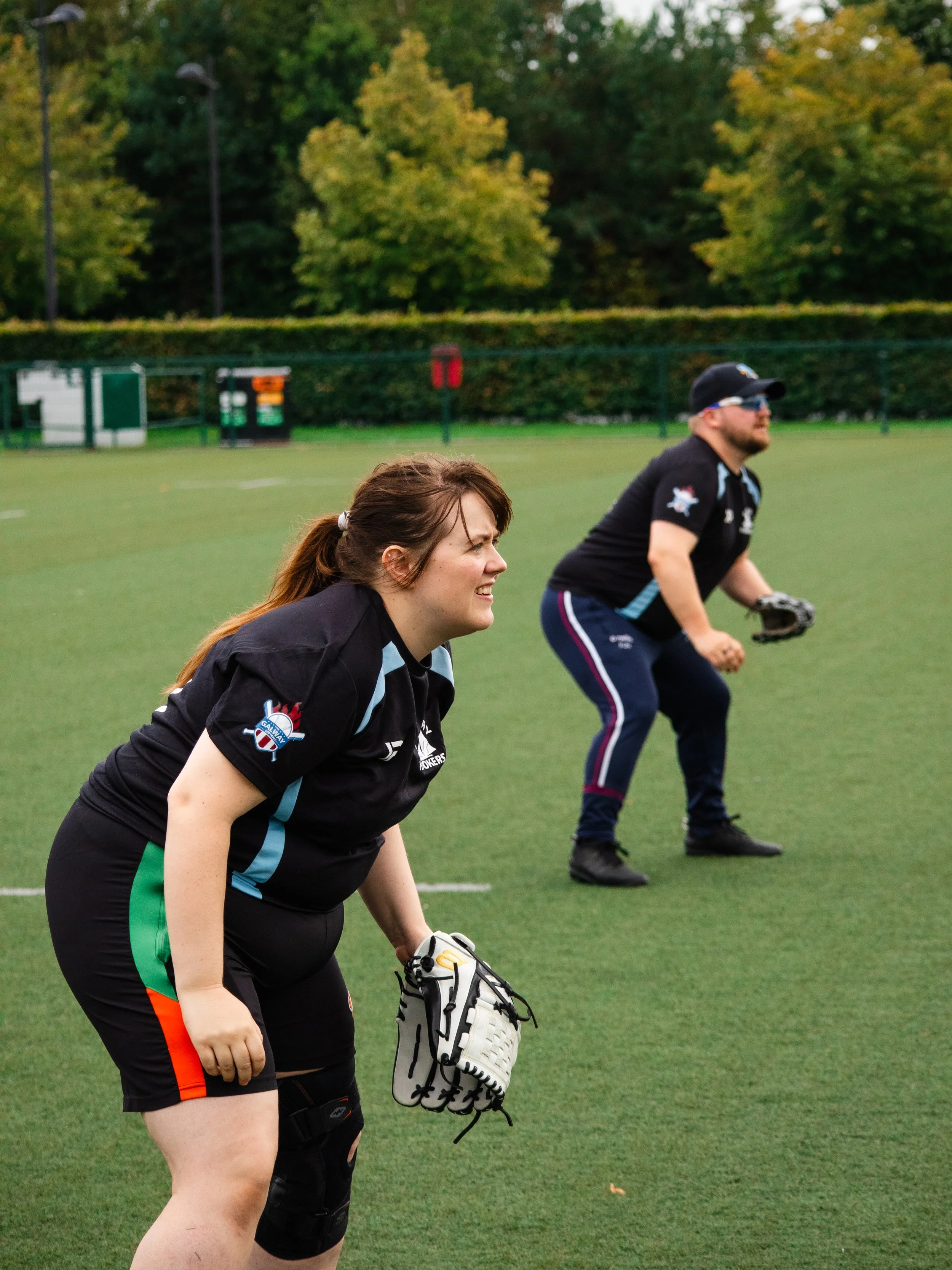 Two adults playing catcher and pitcher in a baseball or softball game on a grass field with trees in the background.