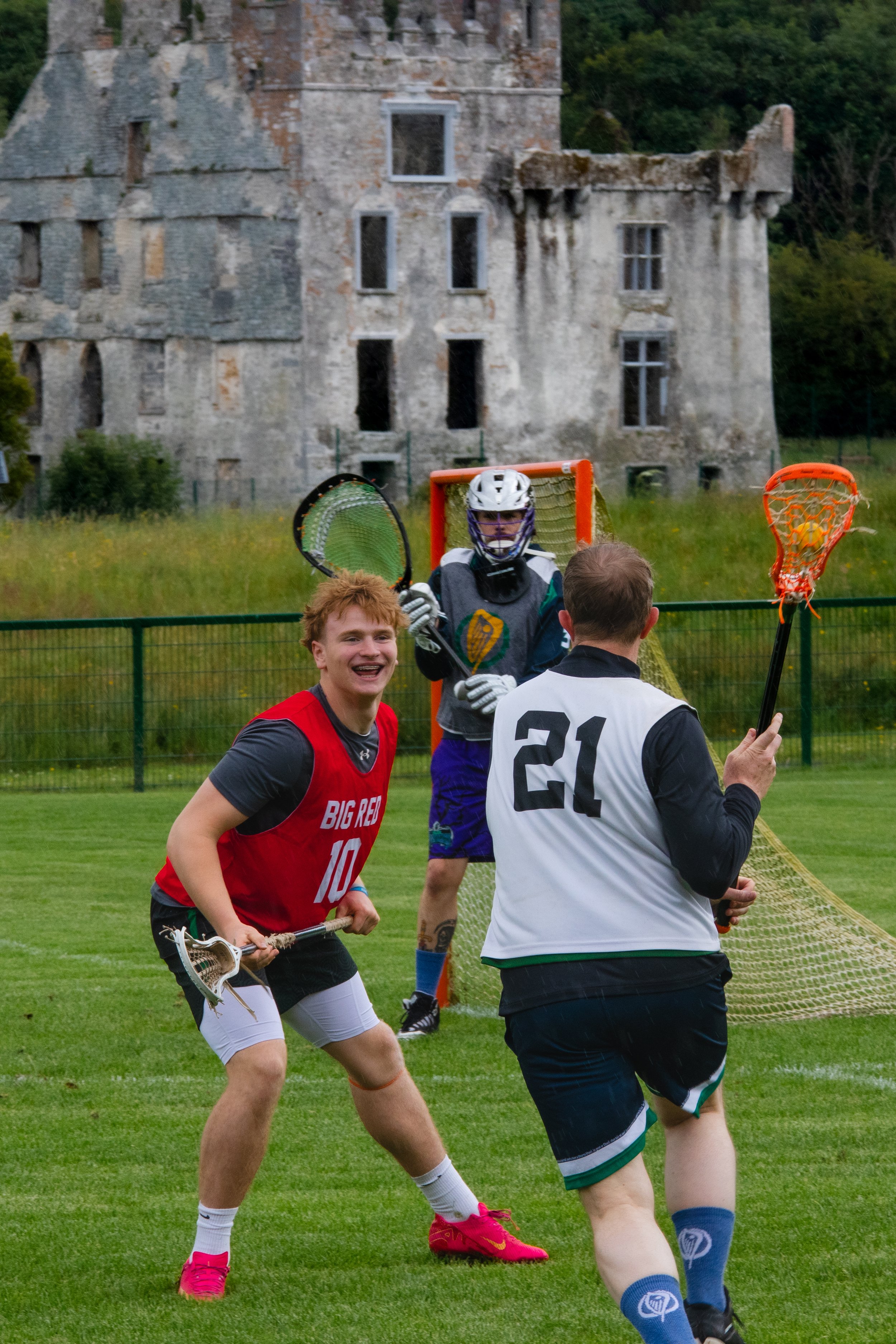 Three young men playing lacrosse on a grassy field with a large, abandoned stone building in the background. One of them is wearing a red jersey, smiling, and holding a lacrosse stick. Another is wearing a black and white jersey with the number 21, h