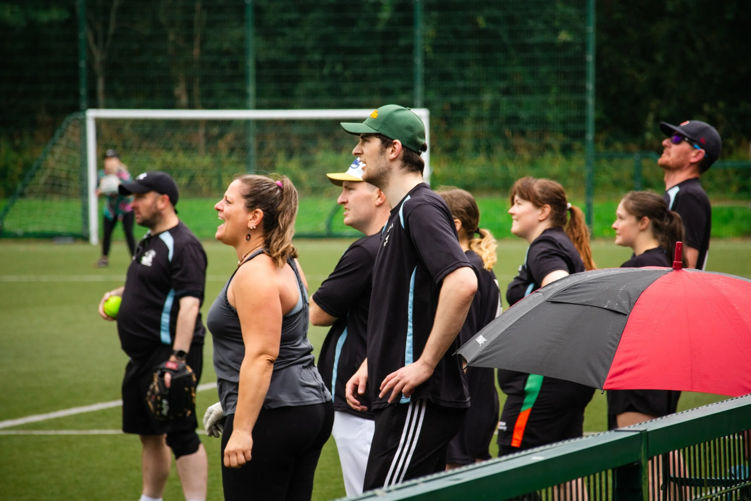 Group of people standing on a grassy sports field, some holding tennis balls, with a soccer goal in the background, and a person holding a red and black umbrella in the foreground.