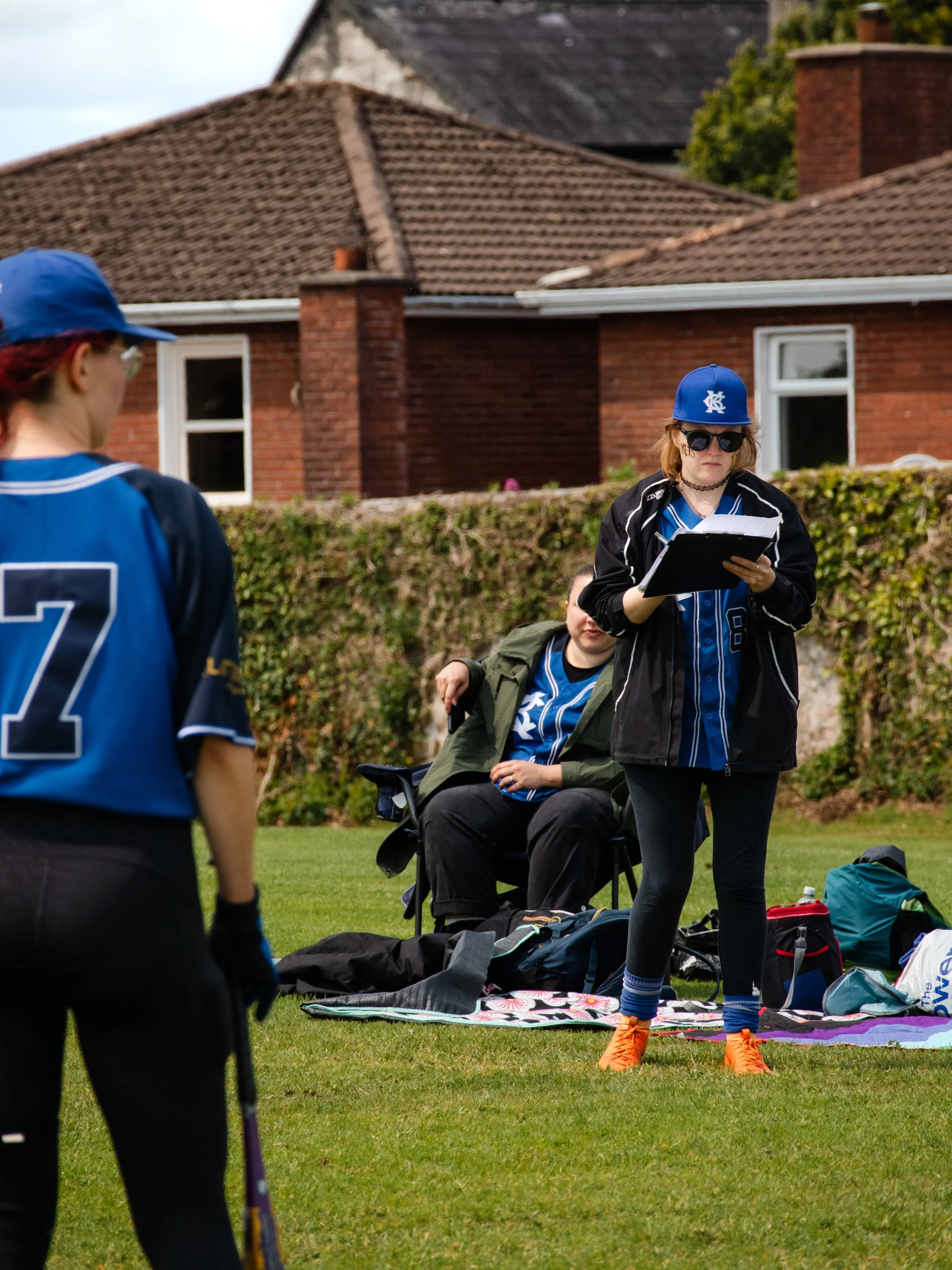 A girl dressed as a baseball player in a blue cap, sunglasses, black jacket, and orange cleats reads from a clipboard during a game or practice, with other players, a coach, and gear visible on the field.