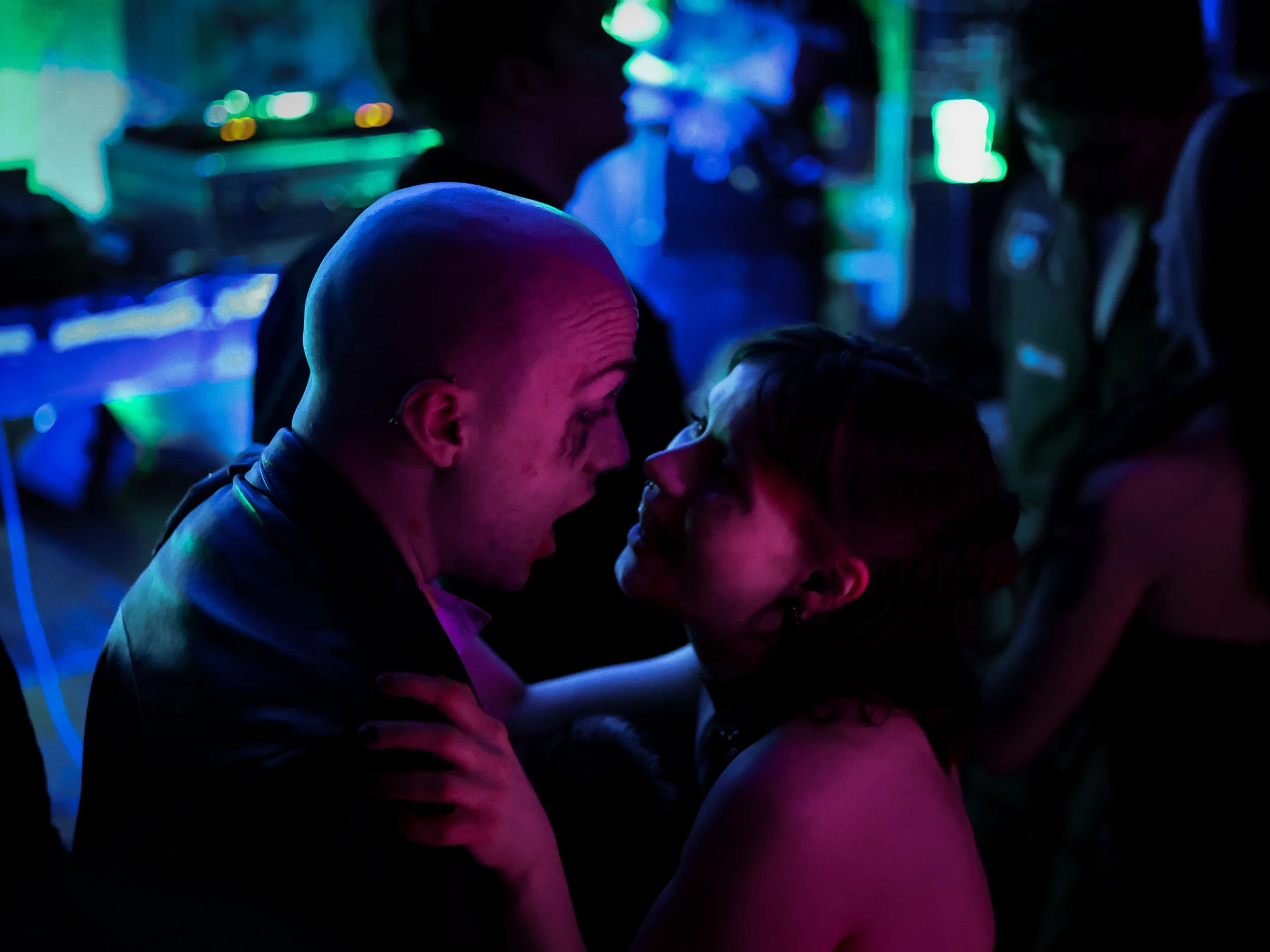 A couple is dancing closely at a party or club with colorful neon lighting, with other people in the background.
